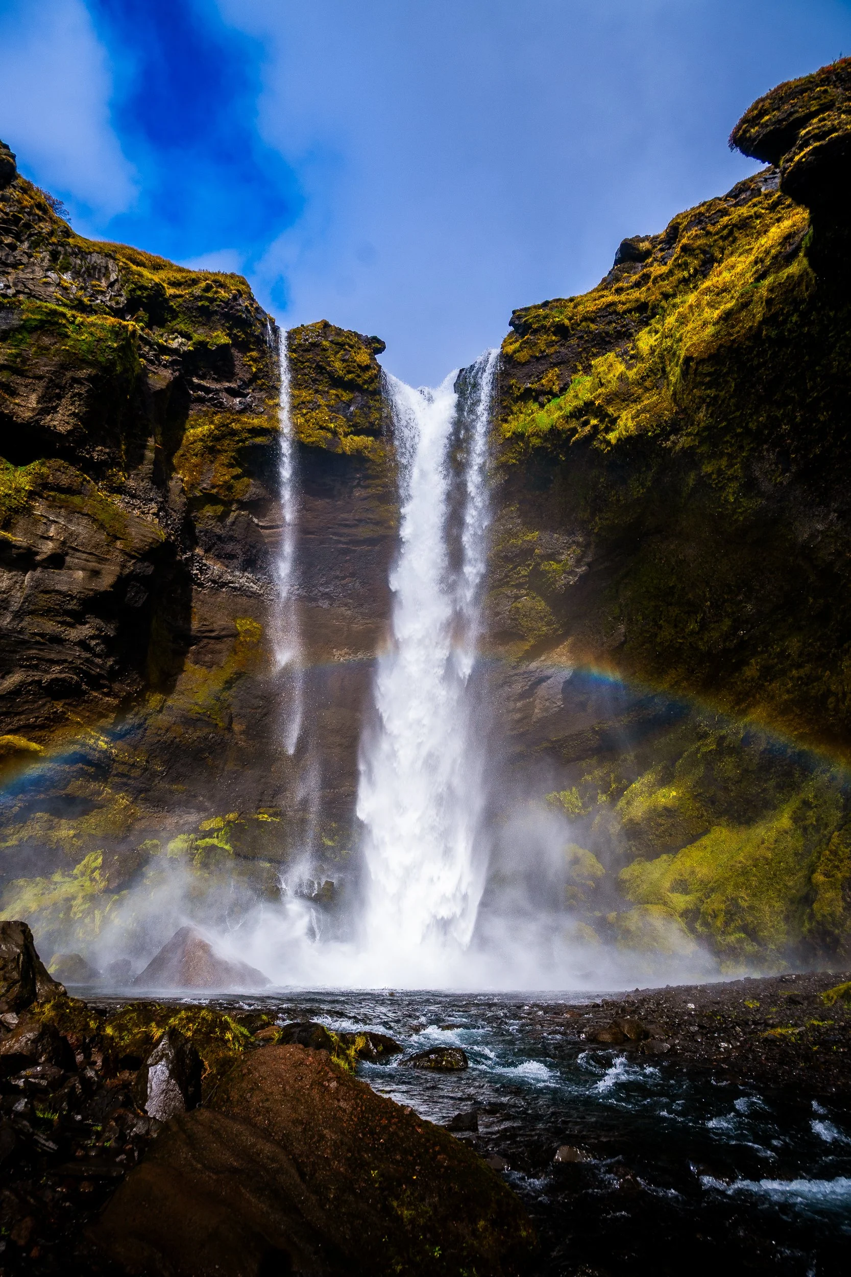 Powerful waterfall cascading through moss-covered volcanic cliffs into a rocky stream beneath a blue sky