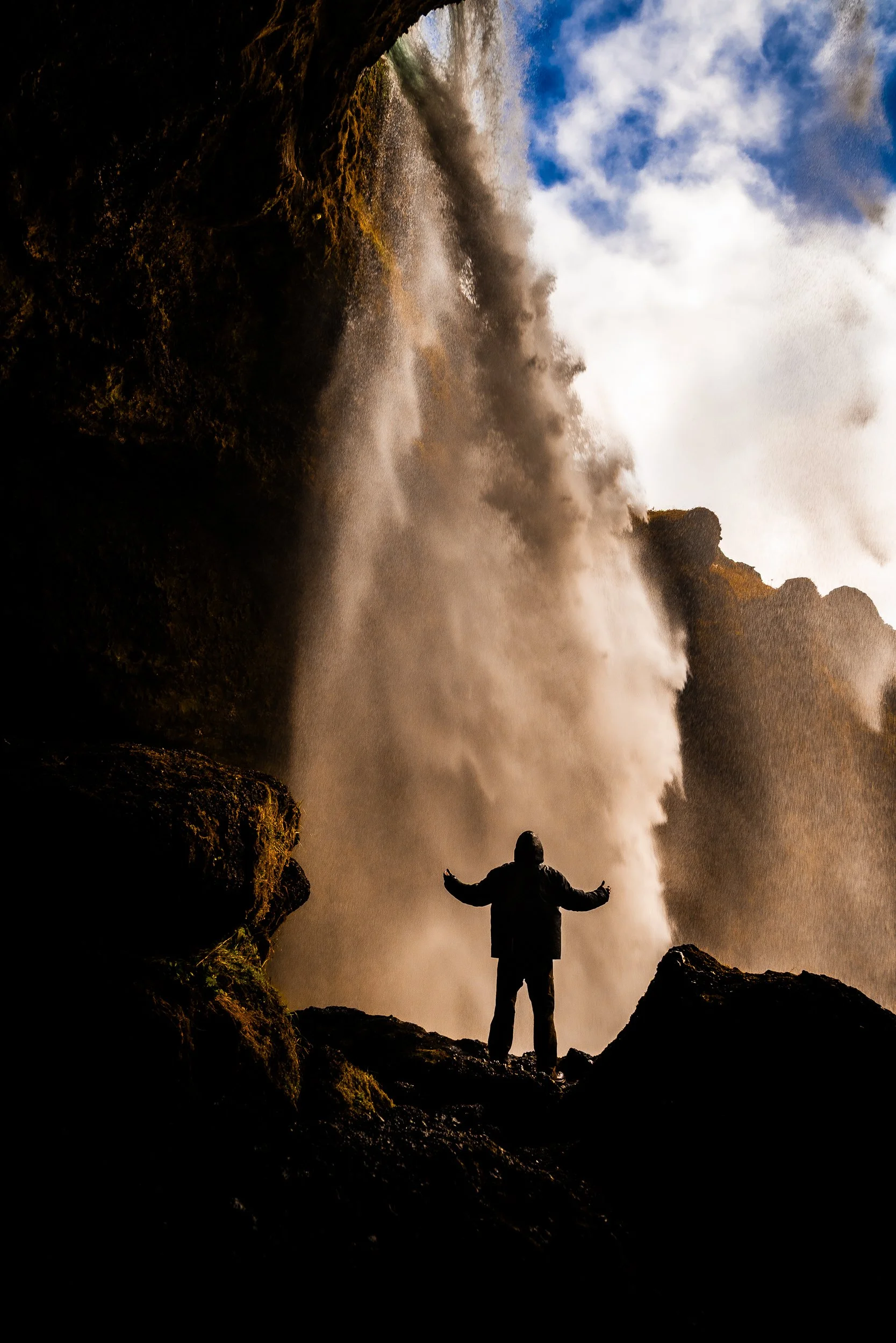 Silhouetted person with arms outstretched beneath a sunlit waterfall surrounded by rocky cliffs