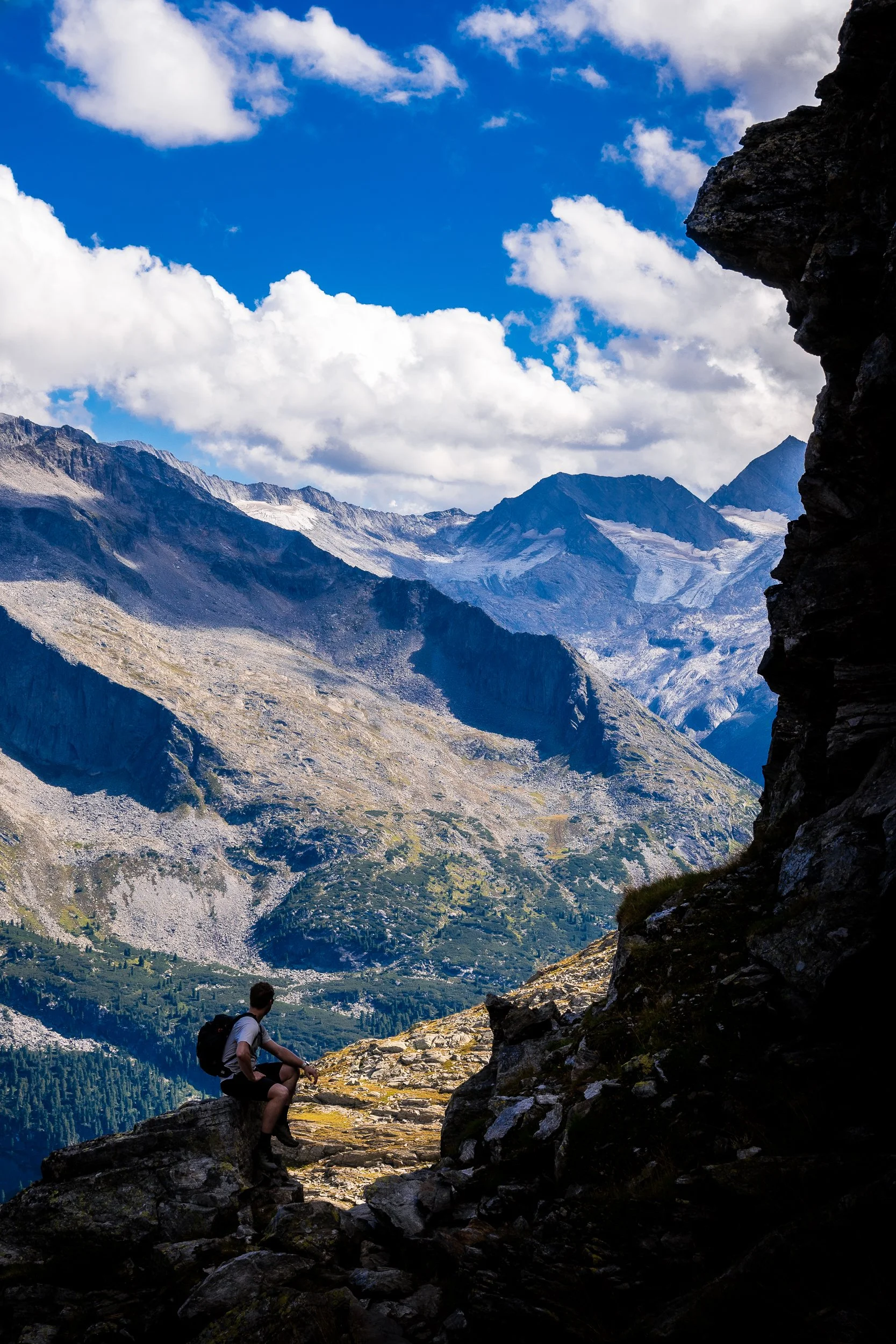 Hiker sitting on a rocky mountain ledge overlooking rugged alpine peaks and glaciers beneath a blue, cloud-filled sky