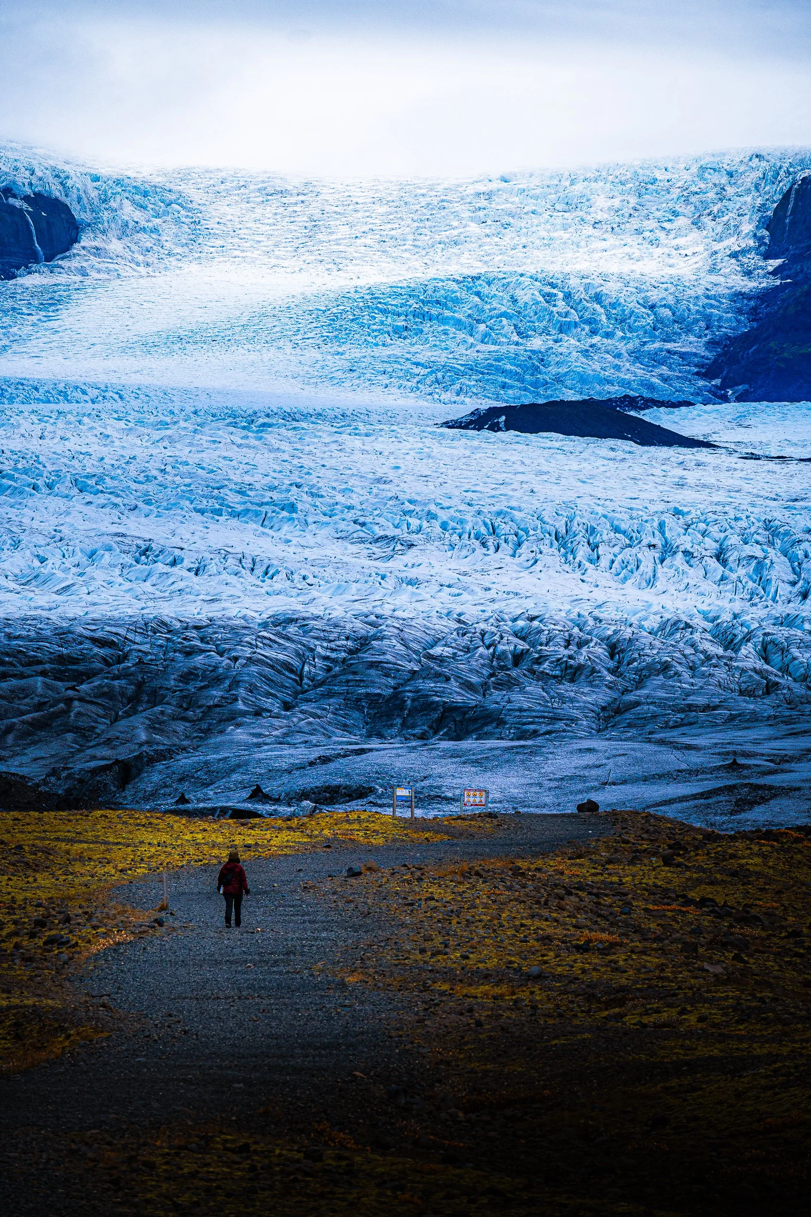 Traveler standing before a vast blue glacier surrounded by volcanic rock and moss-covered terrain