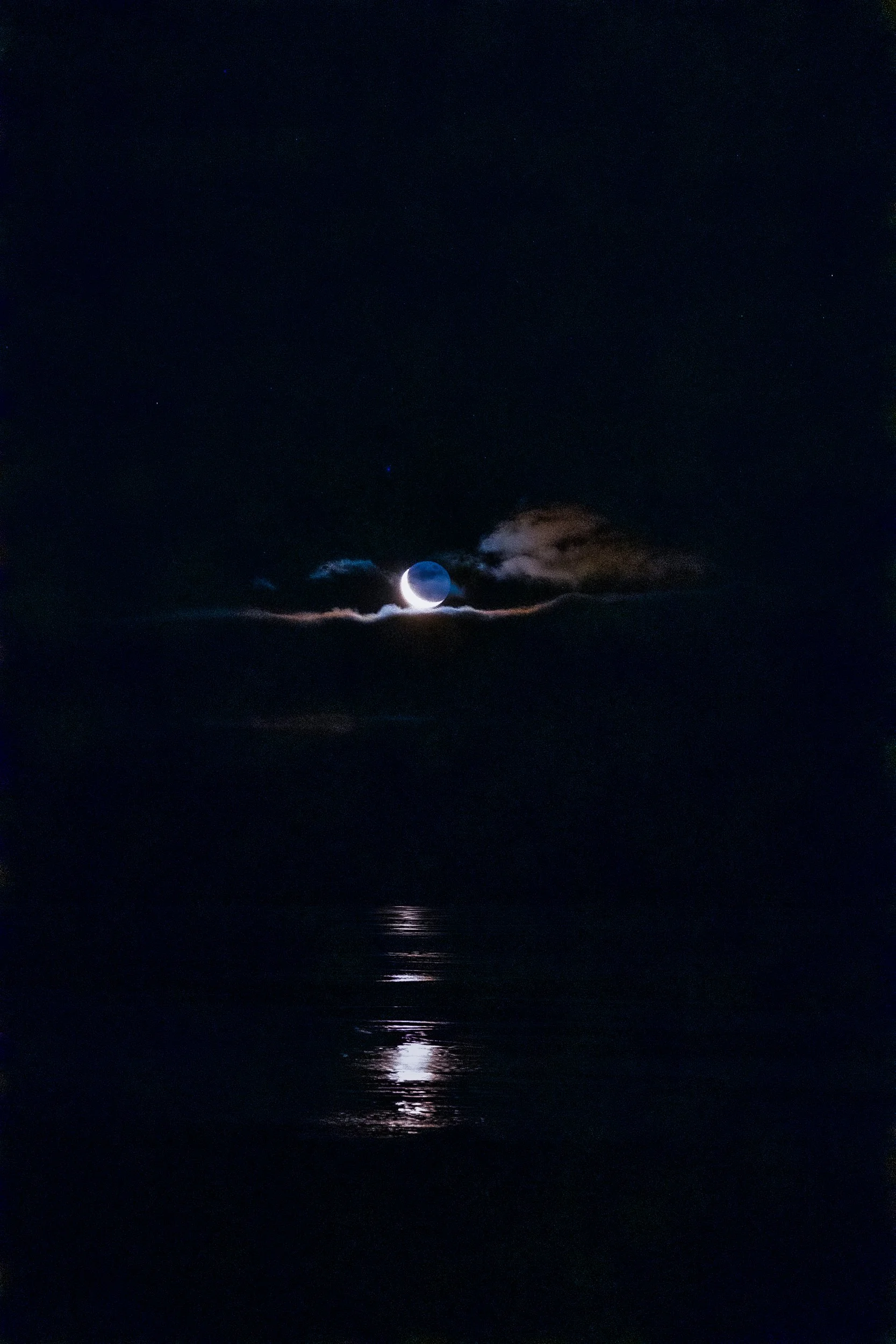 Crescent moon glowing through clouds above dark water, with a shimmering reflection beneath