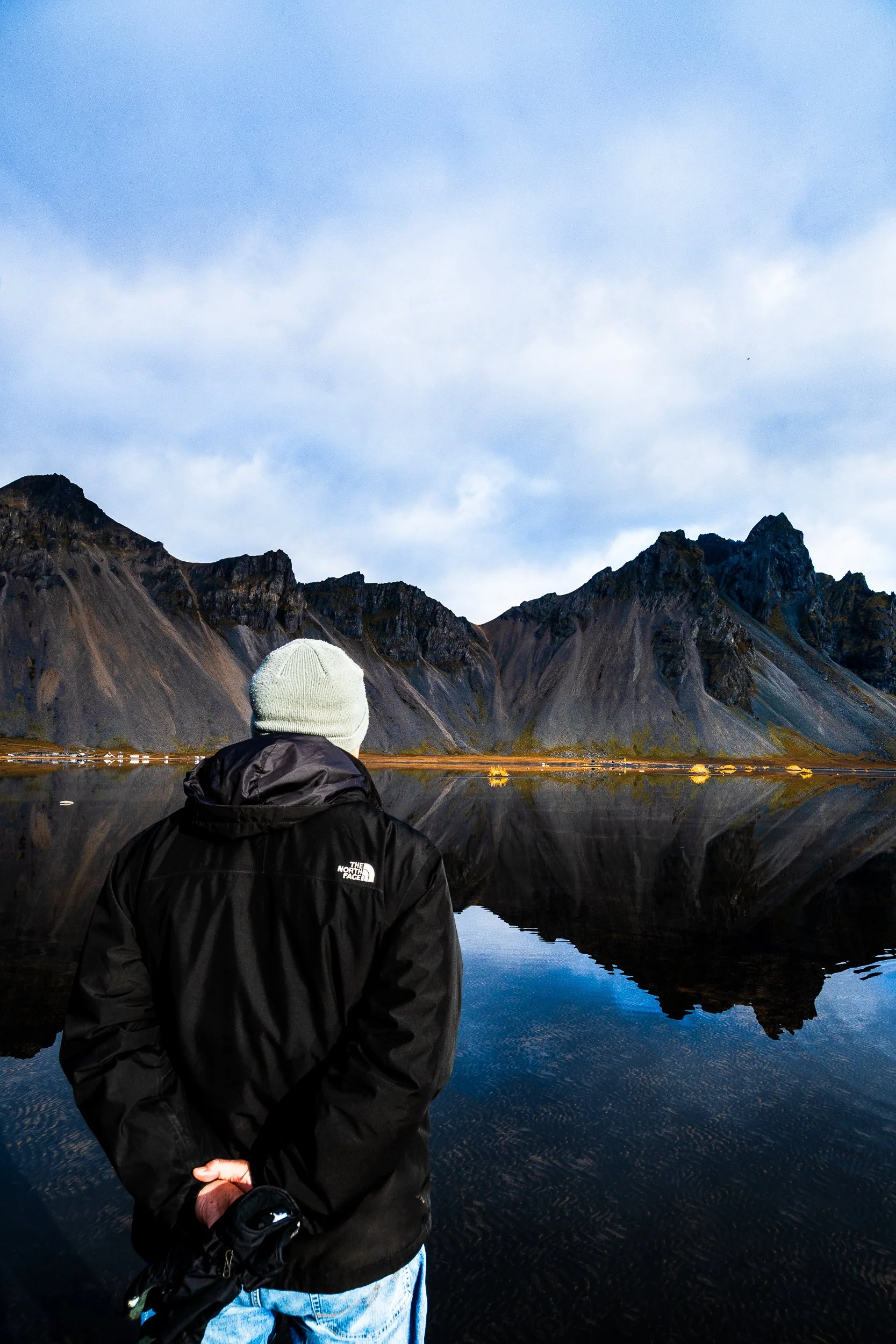 Person in a beanie and black jacket standing beside a reflective mountain lake beneath rugged peaks.