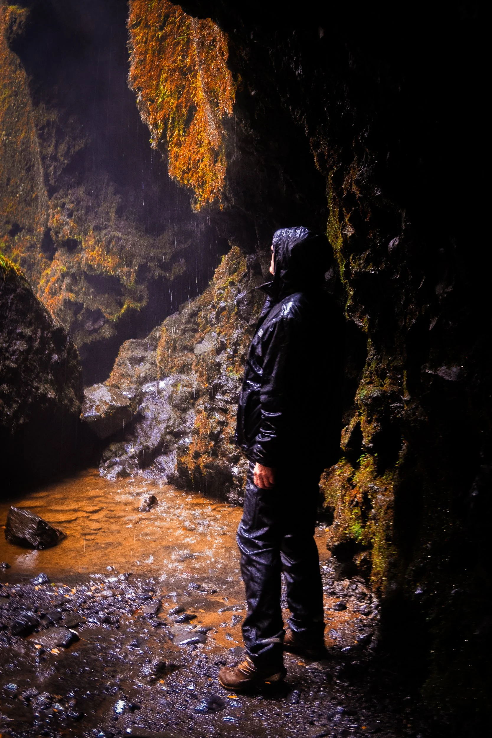 Hiker in black rain gear standing in a moss-covered rocky canyon beside a shallow stream in the rain
