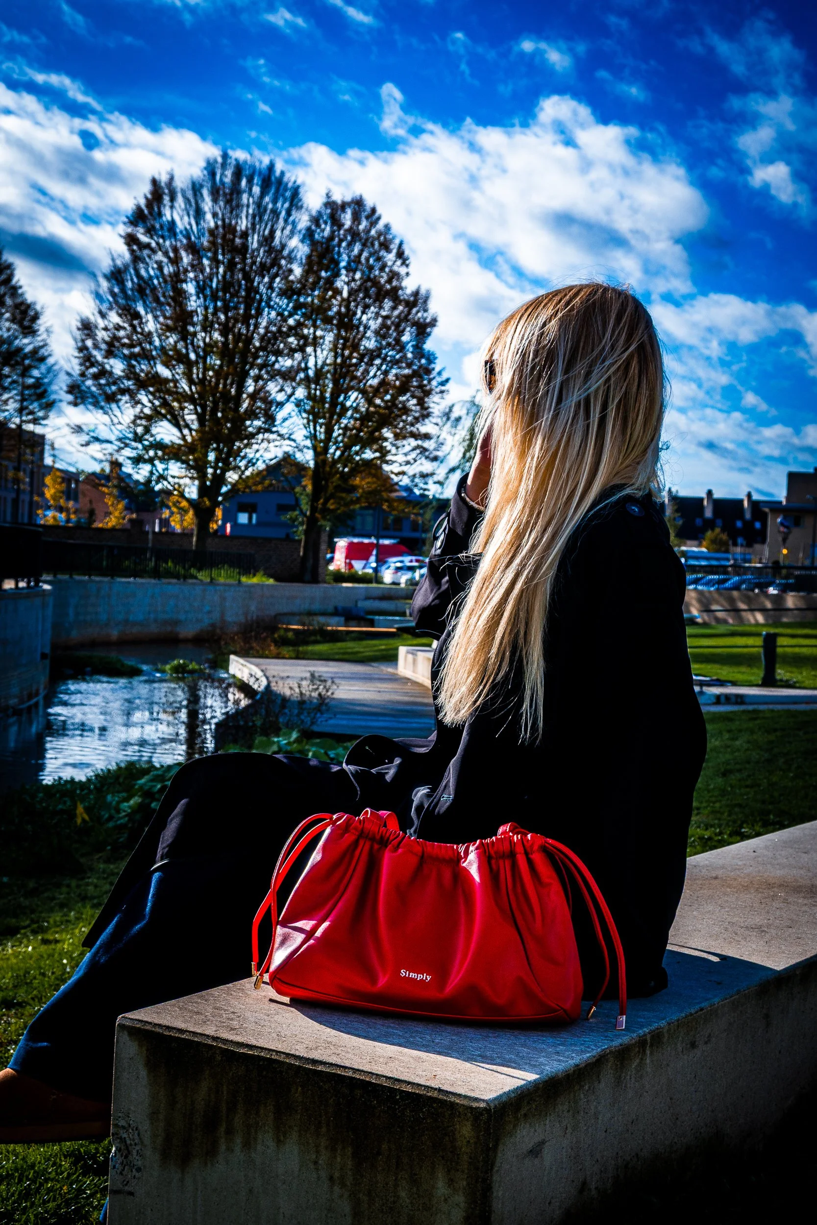 Woman with long blonde hair sitting beside a pond outdoors, with a bright red drawstring bag in the foreground.