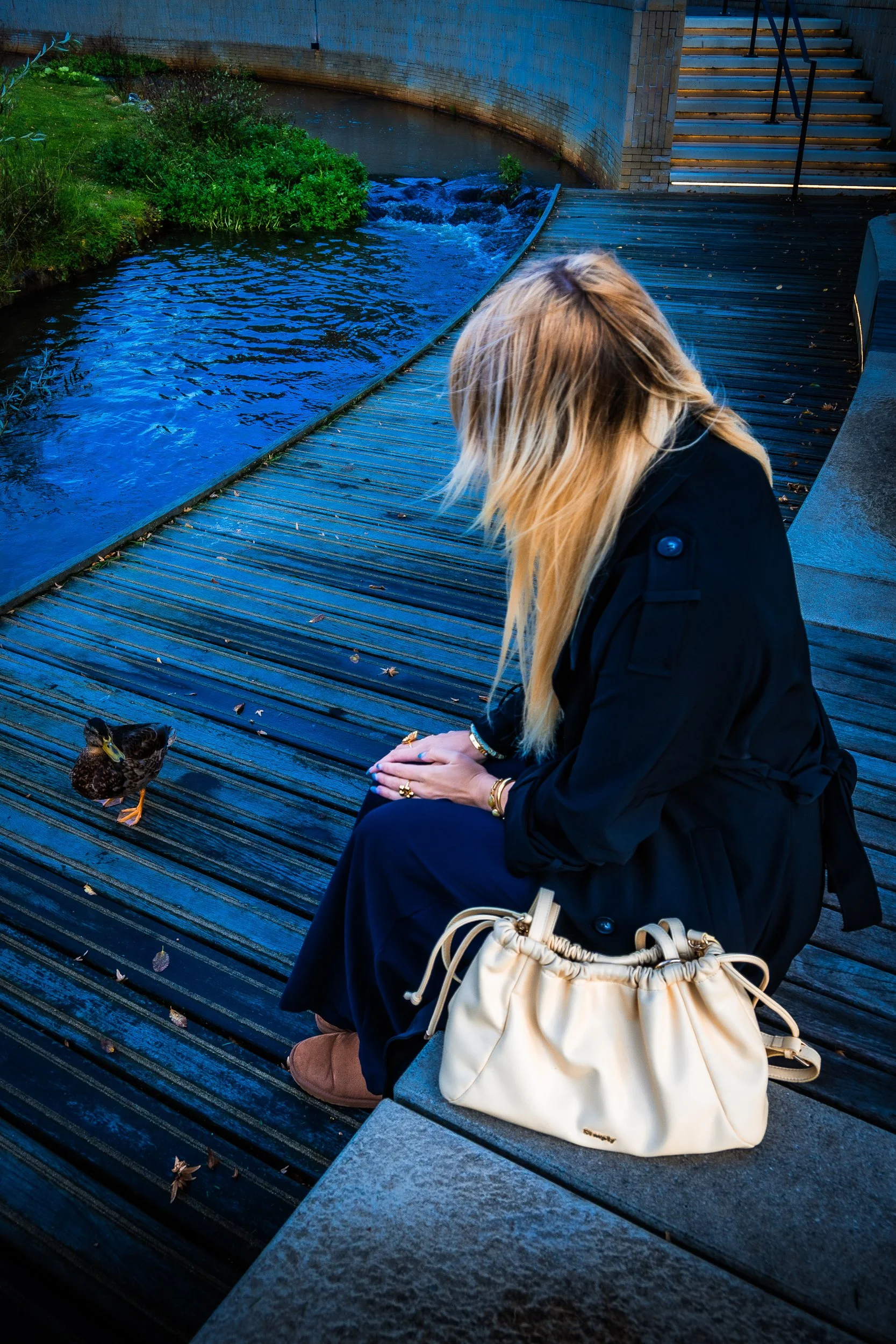 Woman in a black coat sitting on a wooden walkway beside a stream with a duck and stairs in the background