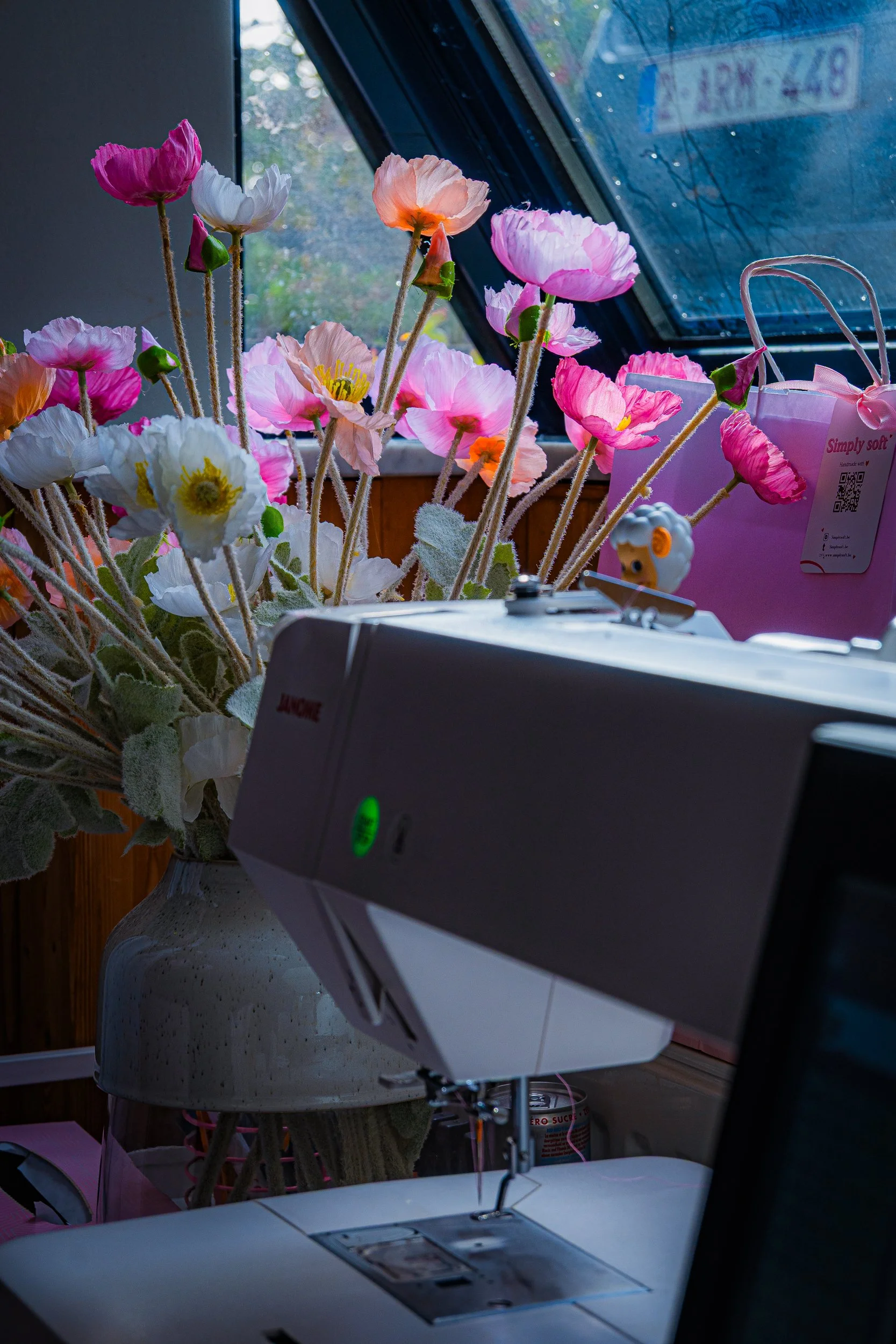 Colorful artificial poppy flowers beside a sewing machine near a sunlit window.