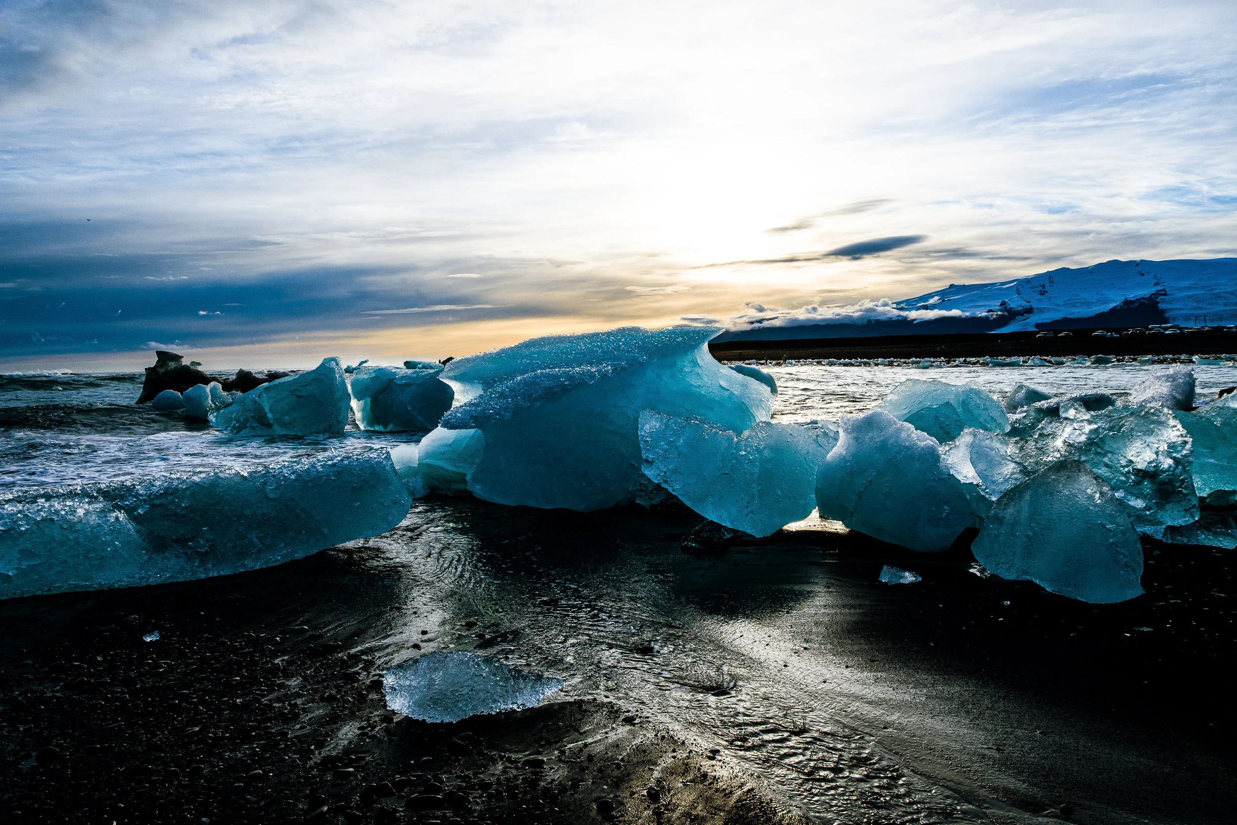 Blue ice chunks scattered along a black sand beach beside a glacial lagoon at sunset, with snow-covered mountains in the background