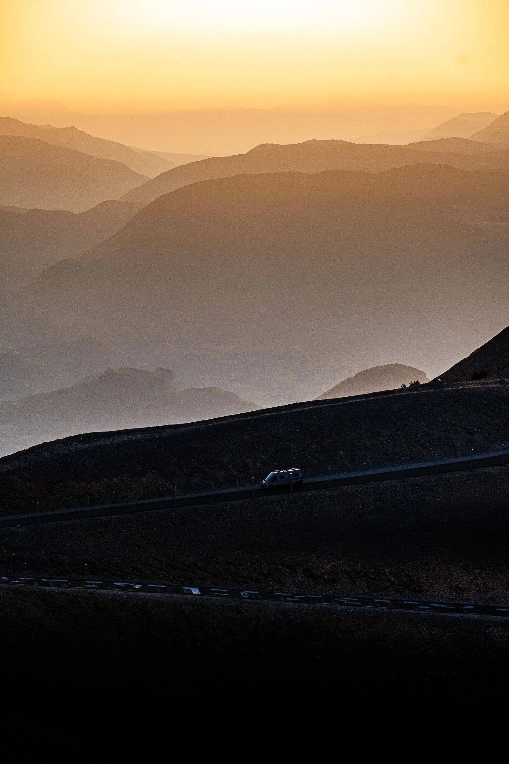 White van traveling along a mountain road above layered mountain ranges at golden hour