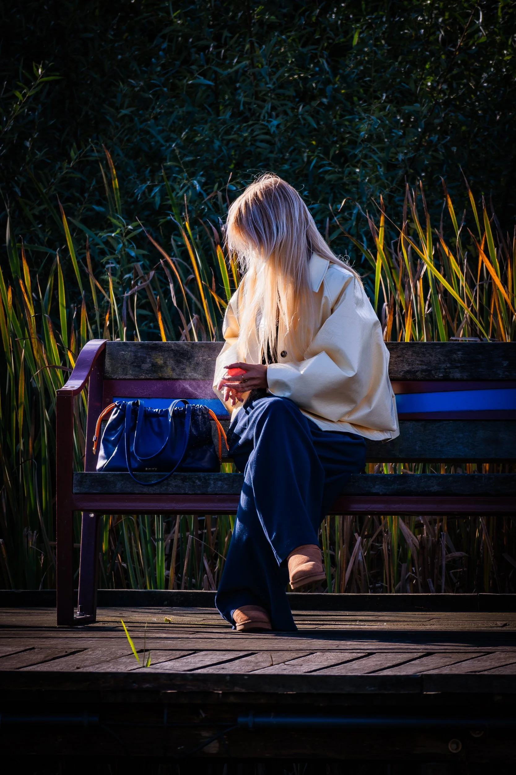 Blonde woman in a white coat sitting on a bench beside a blue handbag in a sunlit marsh.