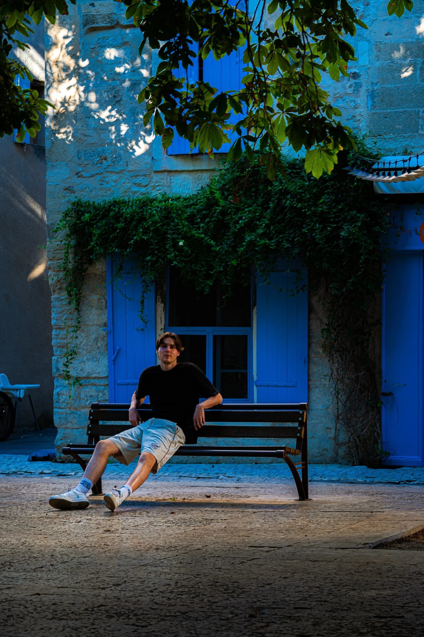 Young man sitting on a bench in front of a blue stone building surrounded by leafy trees