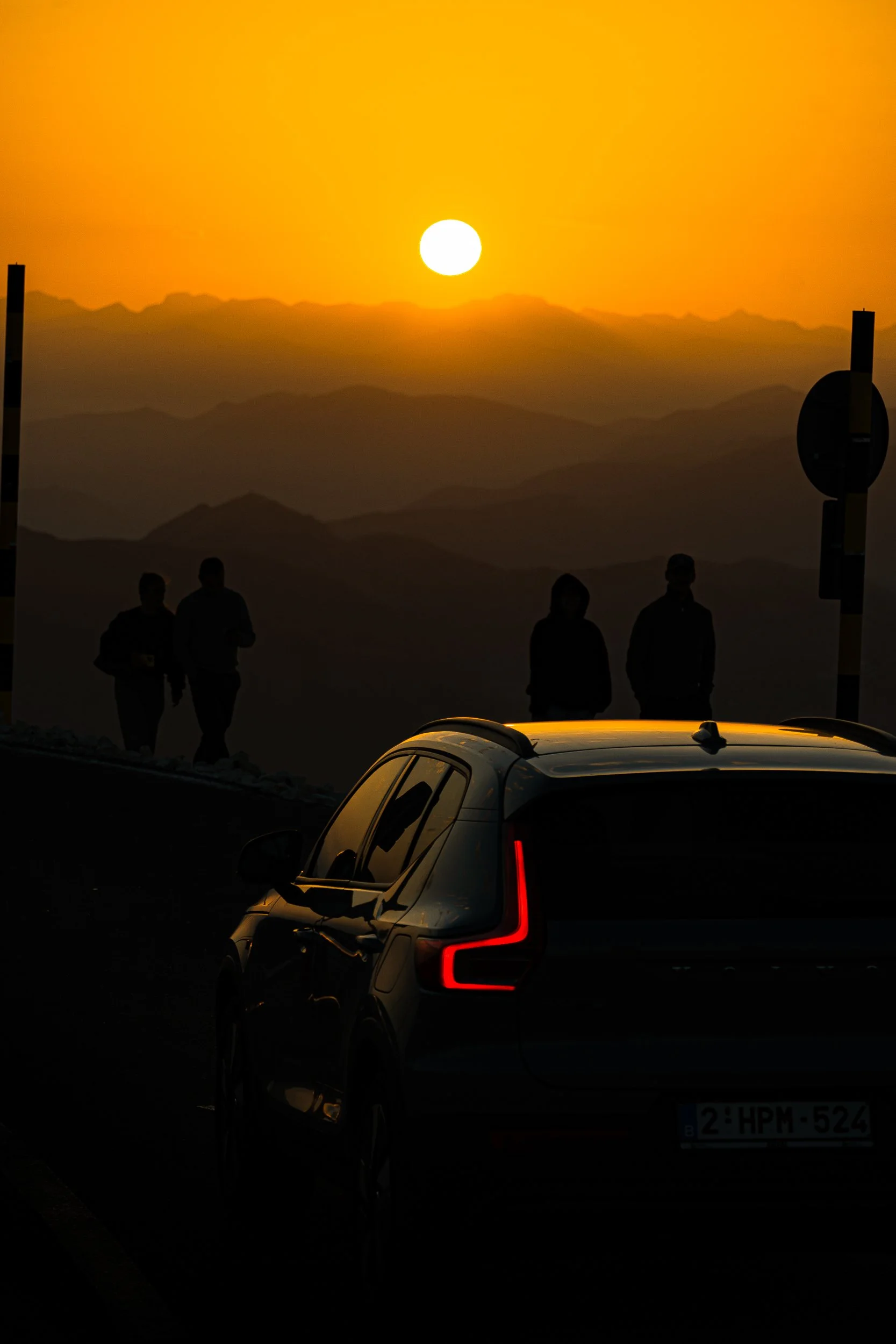 Silhouetted people and a parked car overlooking layered mountains at sunset