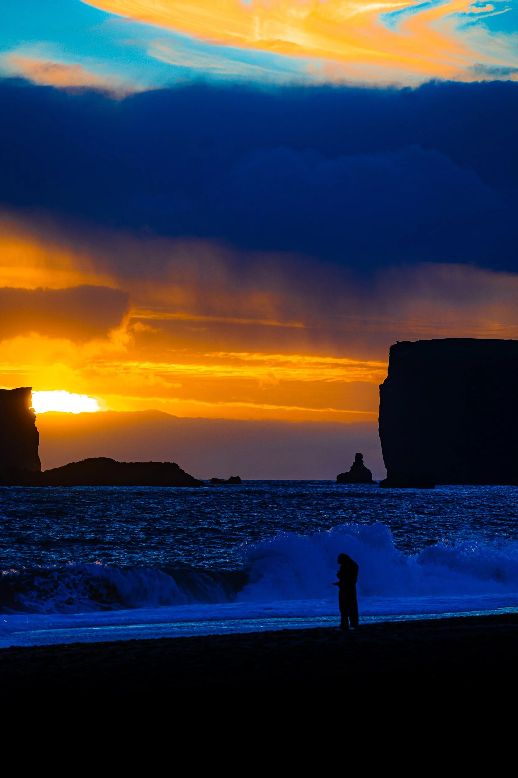 Silhouette of a person watching waves crash against sea cliffs at a vibrant ocean sunset