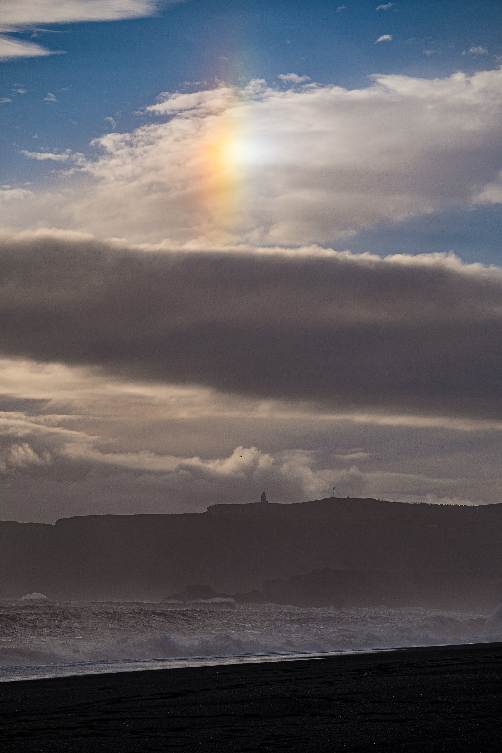 Dramatic sunset over a stormy ocean, with crashing waves and a dark coastal cliff silhouette beneath layered clouds