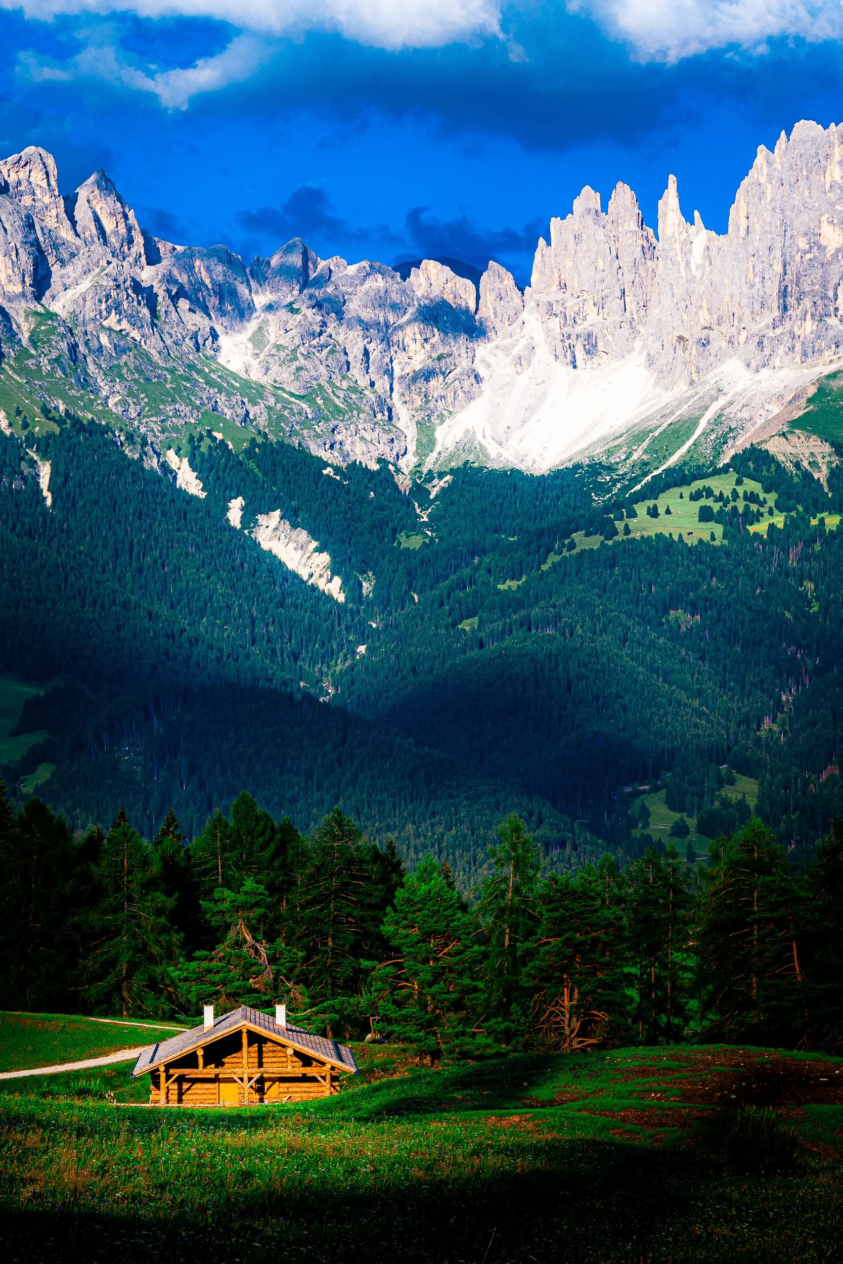 Wooden alpine cabin in a meadow beneath dramatic limestone peaks and dense evergreen forests