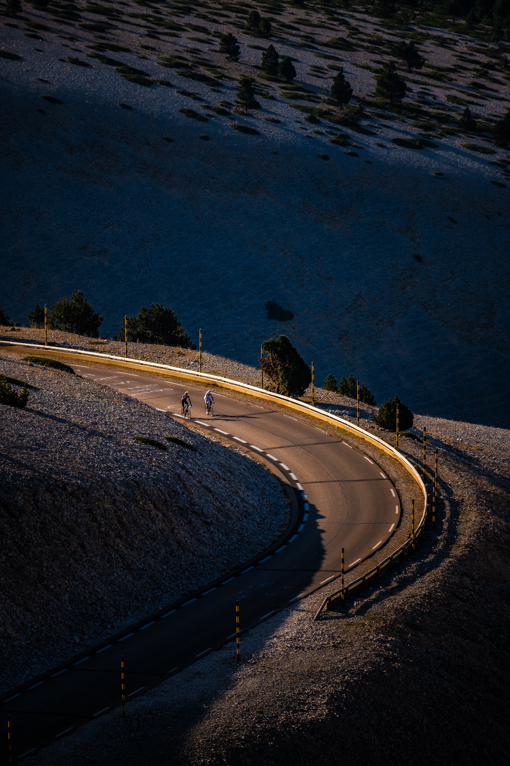 Two cyclists riding along a winding mountain road covered in gravel, with sparse alpine trees and reflective roadside posts.
