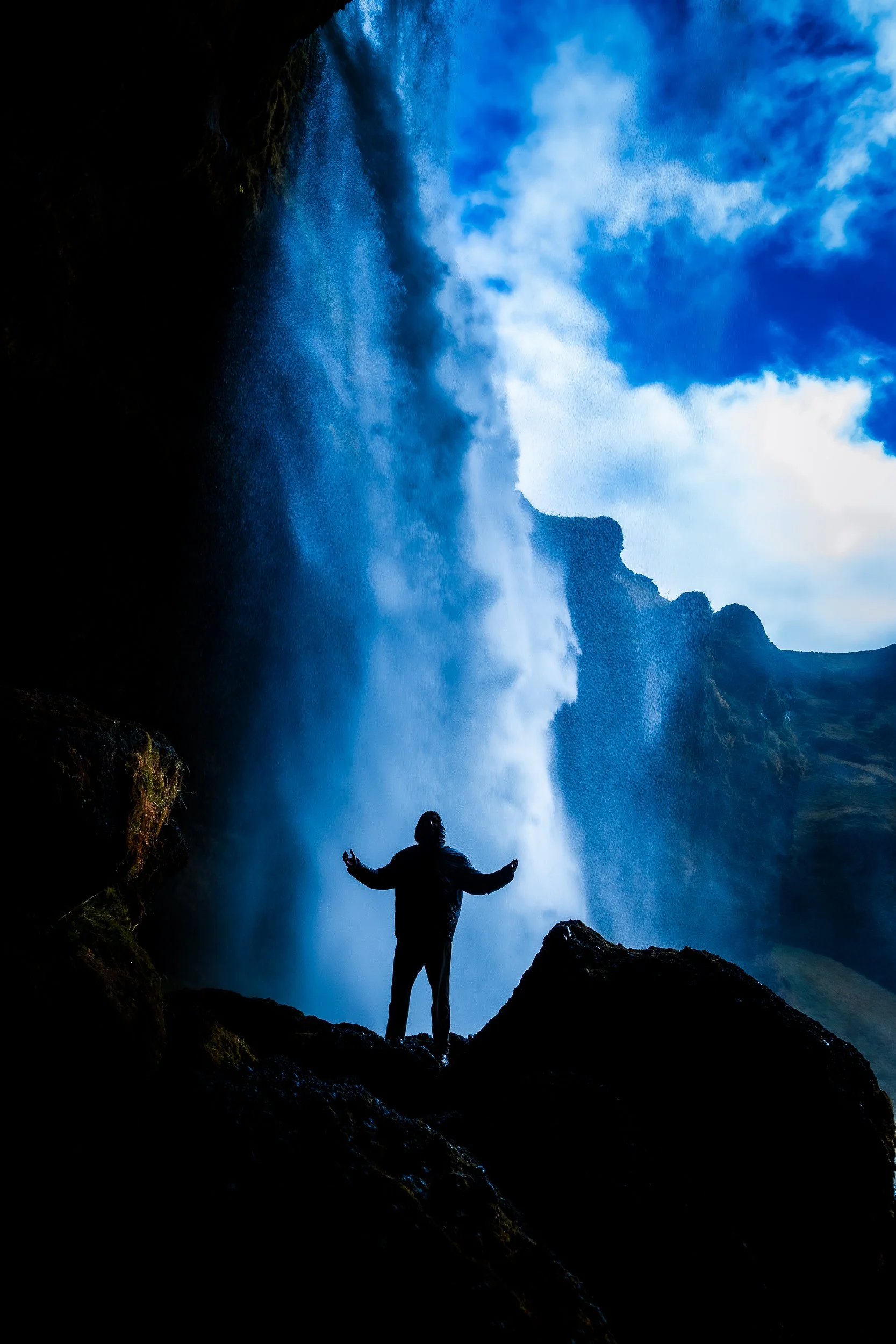 Silhouette of a person standing with arms outstretched beneath a powerful waterfall, surrounded by dark cliffs and blue sky.