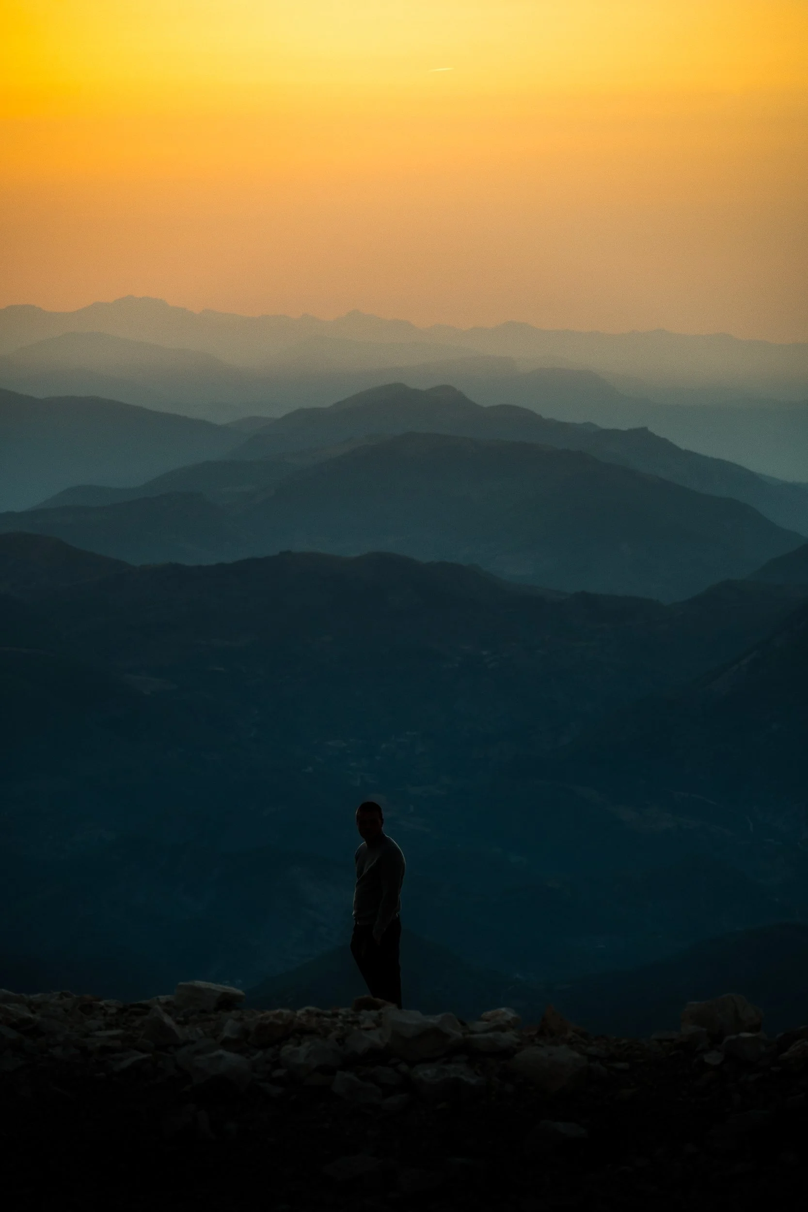 Silhouetted person standing on a rocky mountain ridge overlooking layered mountains at sunset
