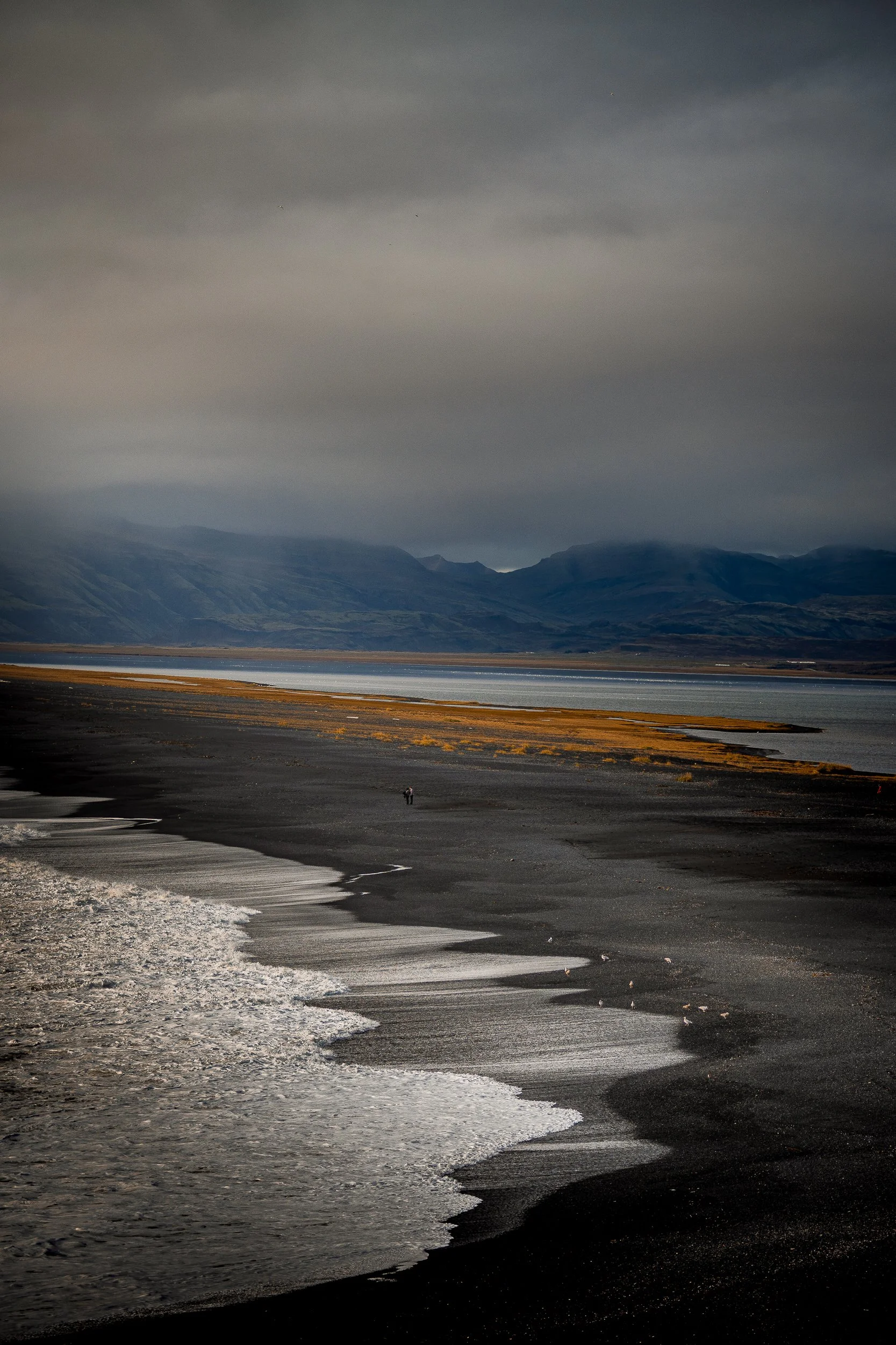 Black sand beach with foamy waves, a calm lagoon, distant mountains, and a lone person beneath a stormy sky