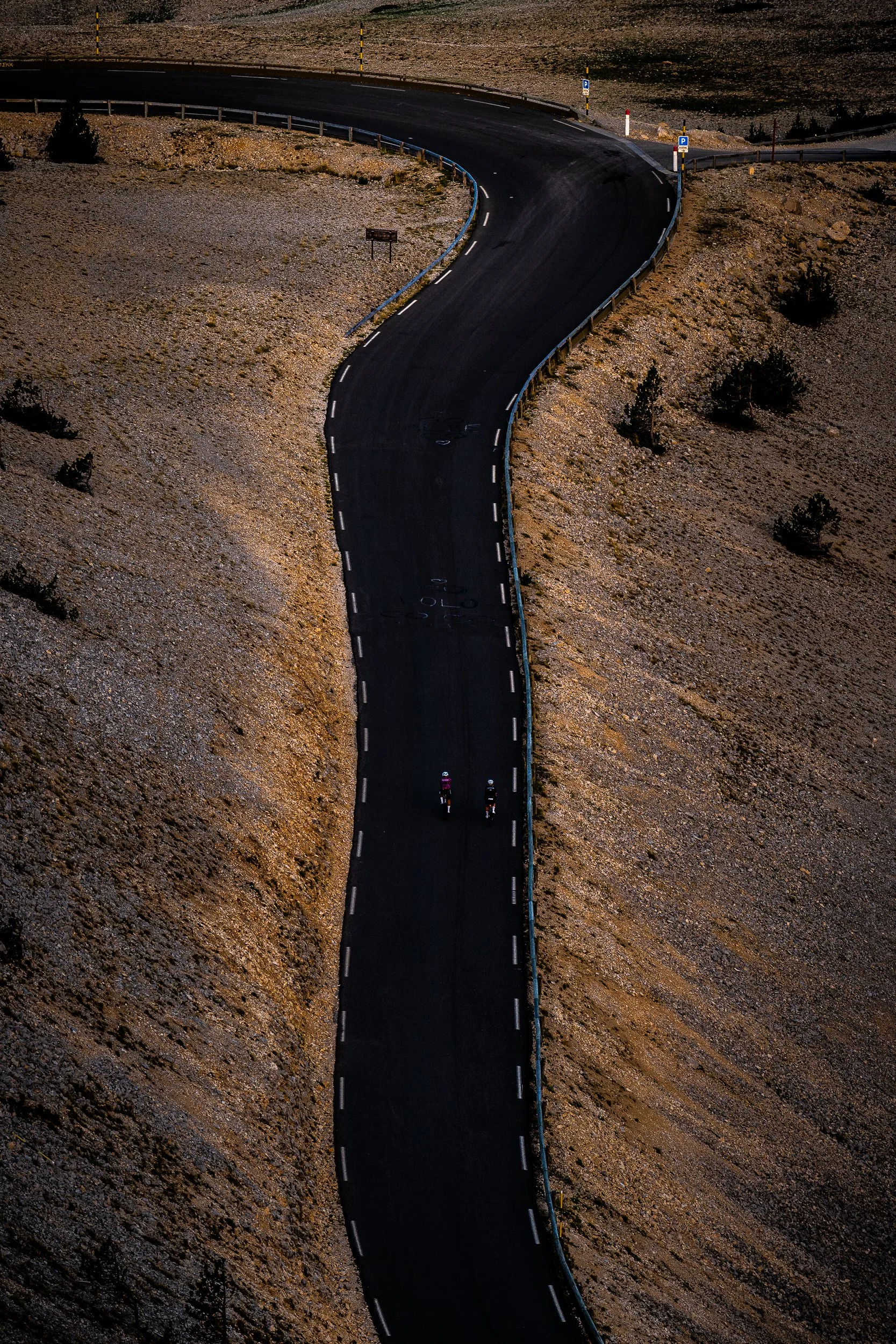 Aerial view of two cyclists riding on a winding mountain road through a rocky landscape.