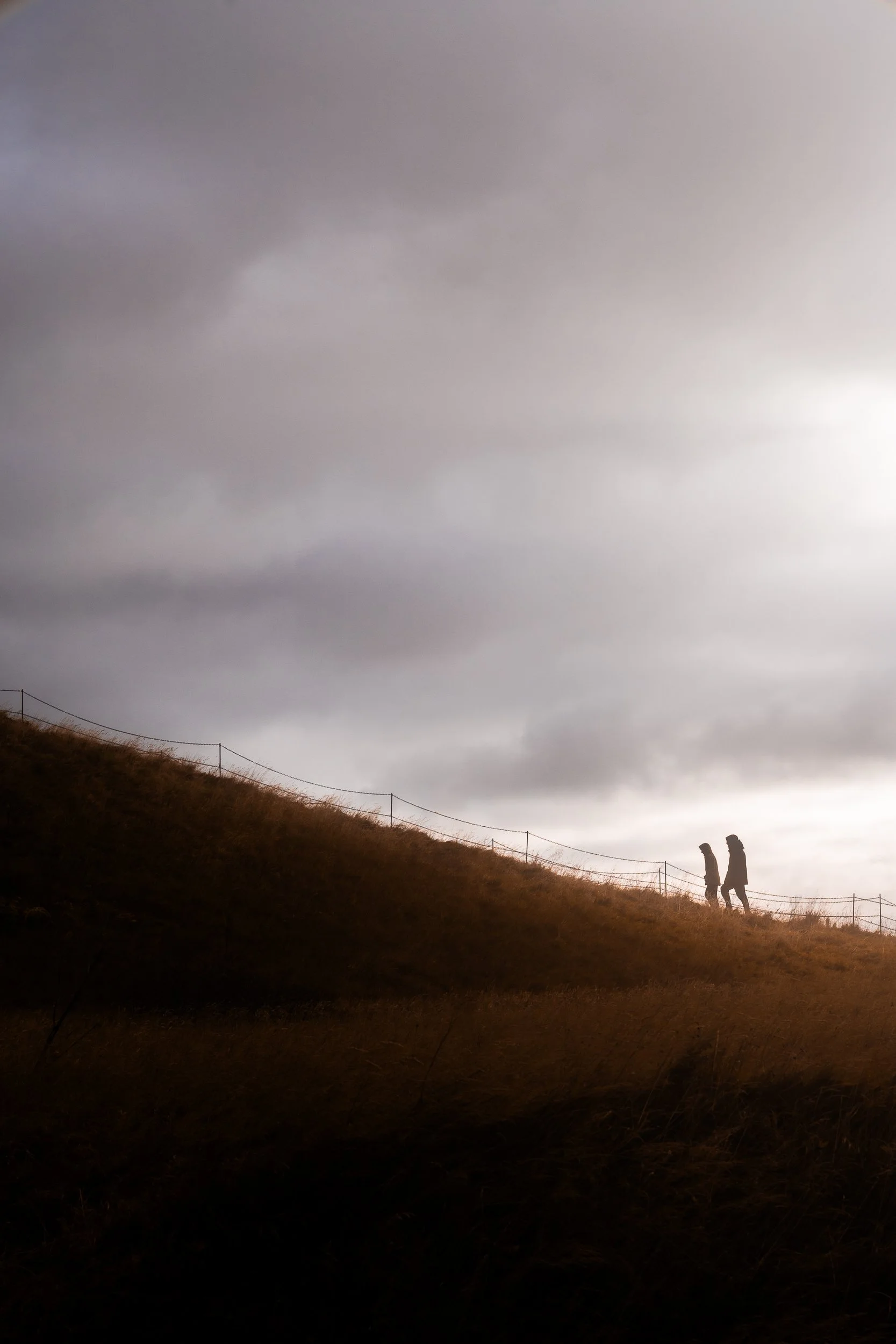 Two silhouetted hikers walking along a grassy hillside beneath an overcast sky