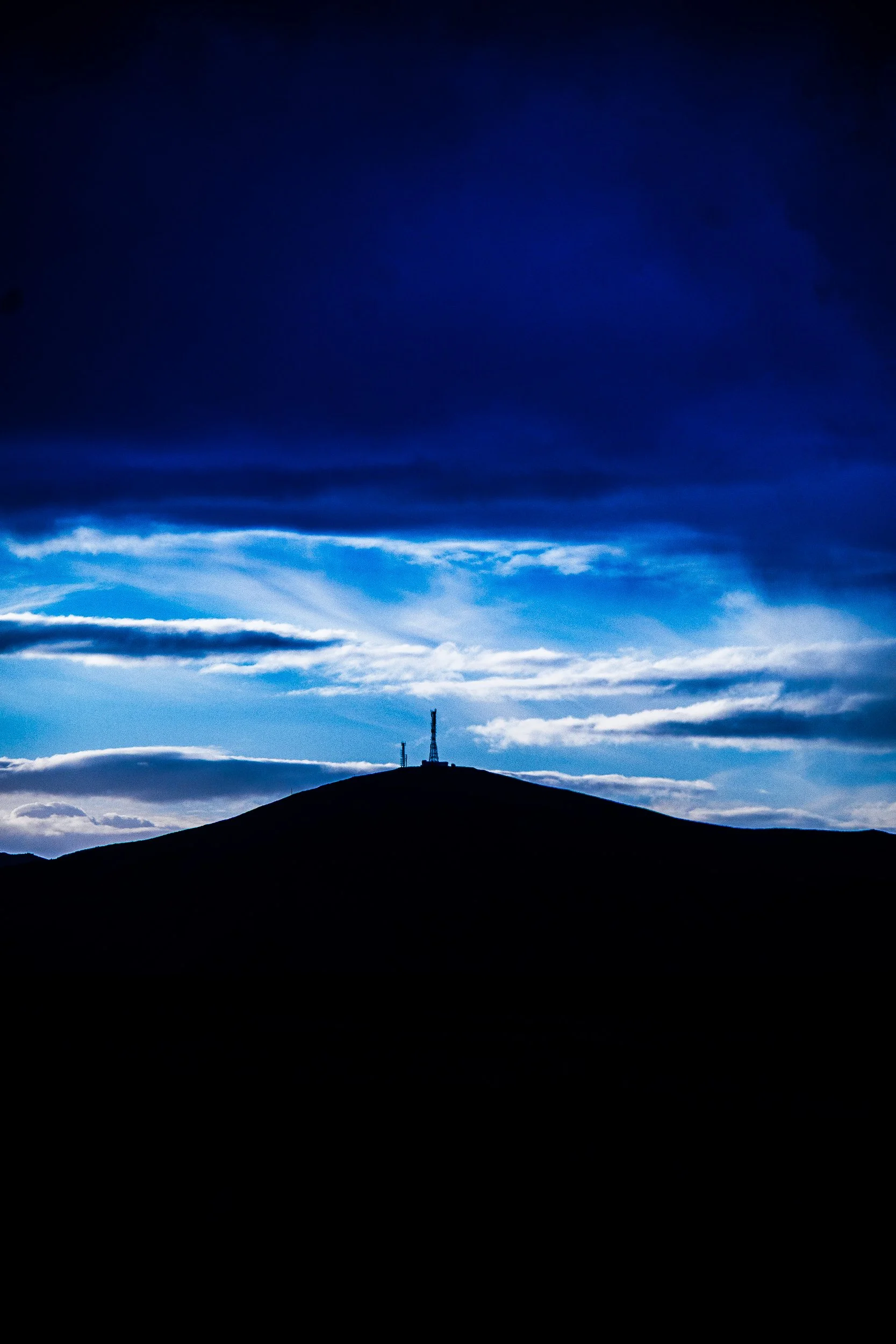 Dark mountain ridge with communication towers silhouetted against a dramatic blue cloud-filled sky