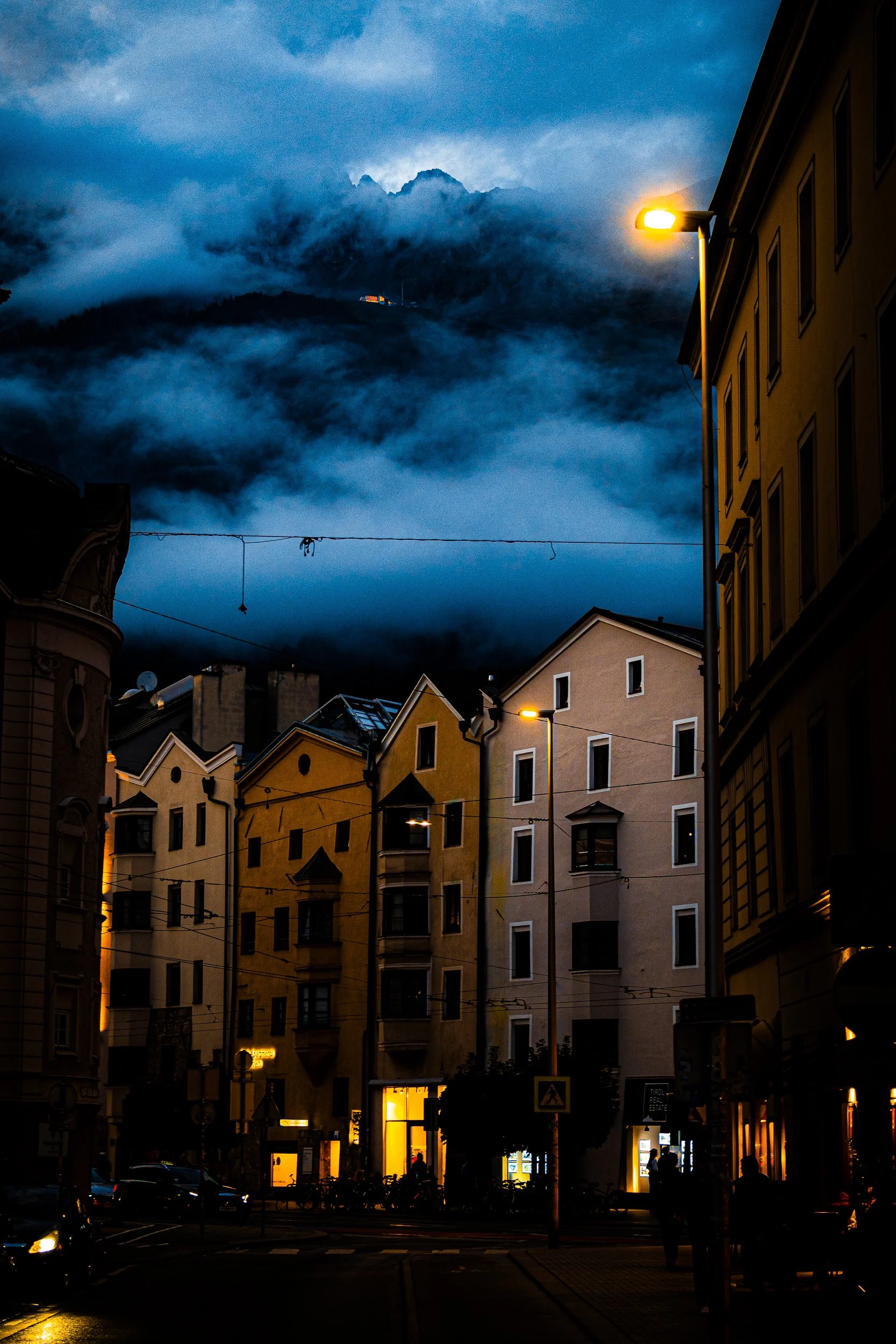 Nighttime city street with historic buildings, glowing streetlights, overhead wires, and dramatic blue storm clouds.