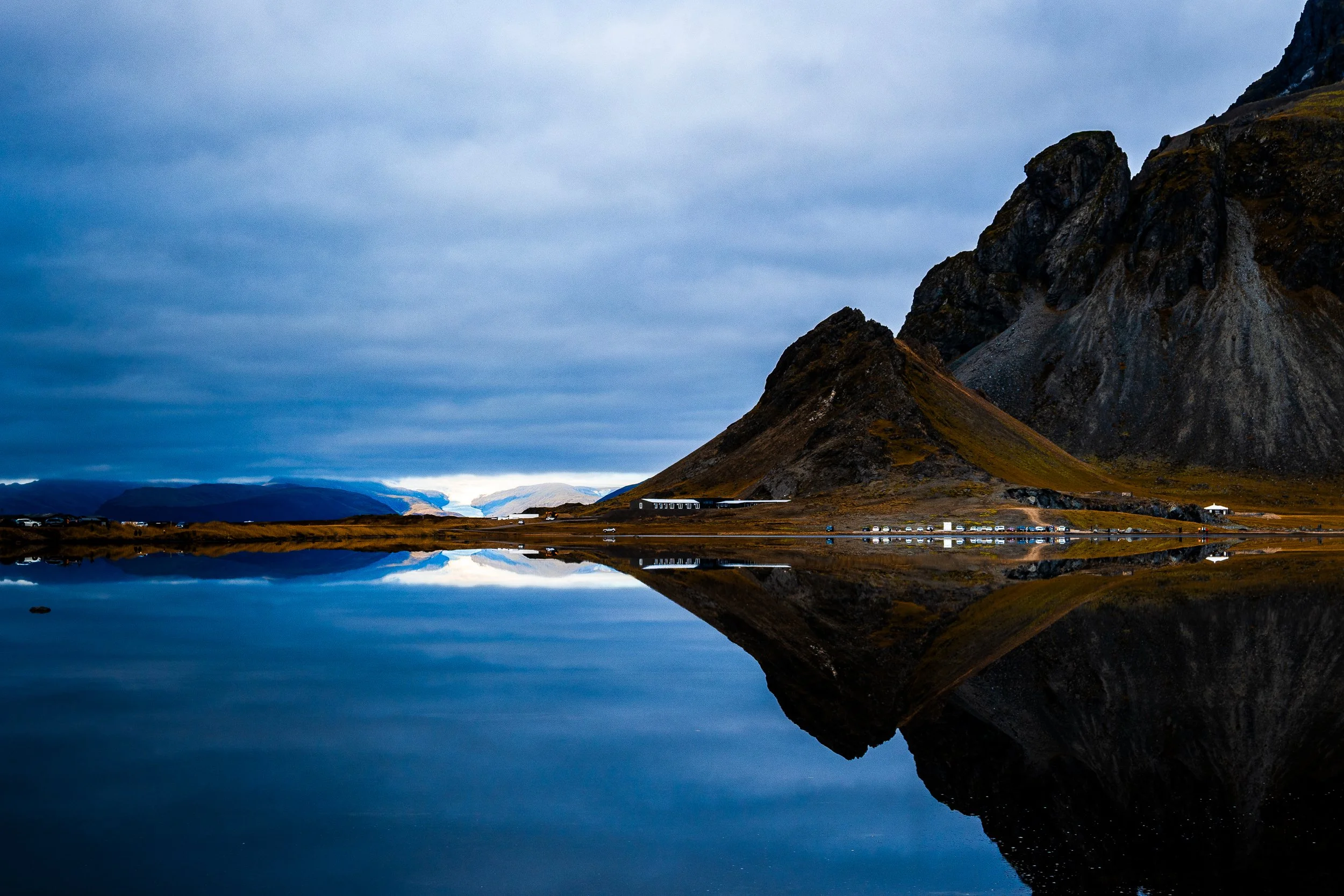 Dramatic Icelandic mountain landscape reflected in a calm lake beneath a cloudy blue sky