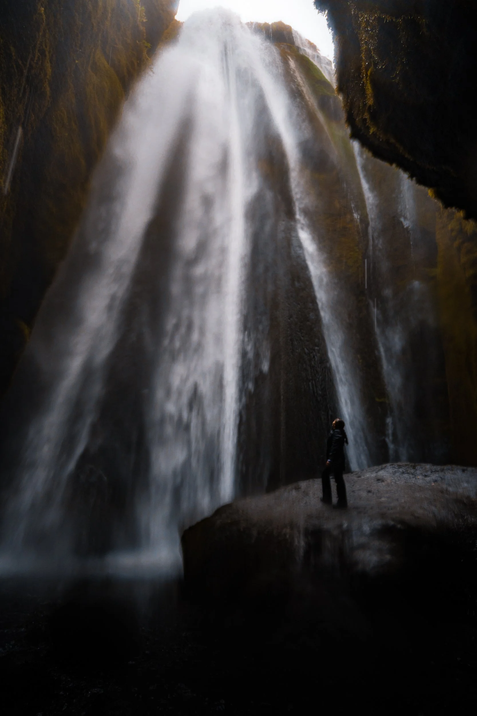 Person standing beneath a towering waterfall surrounded by moss-covered cliffs