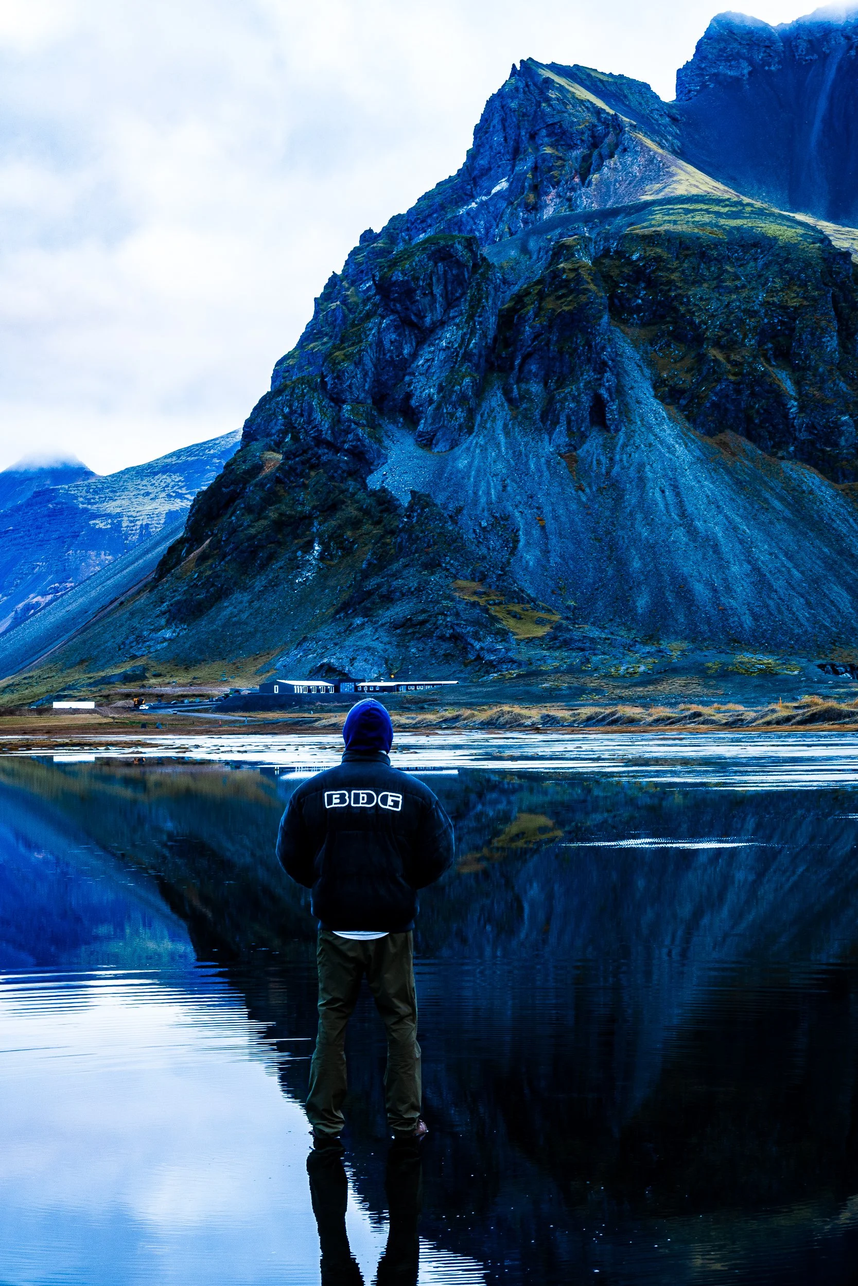 Person in a hooded jacket standing in a reflective mountain lake beneath a rugged, snow-dusted peak