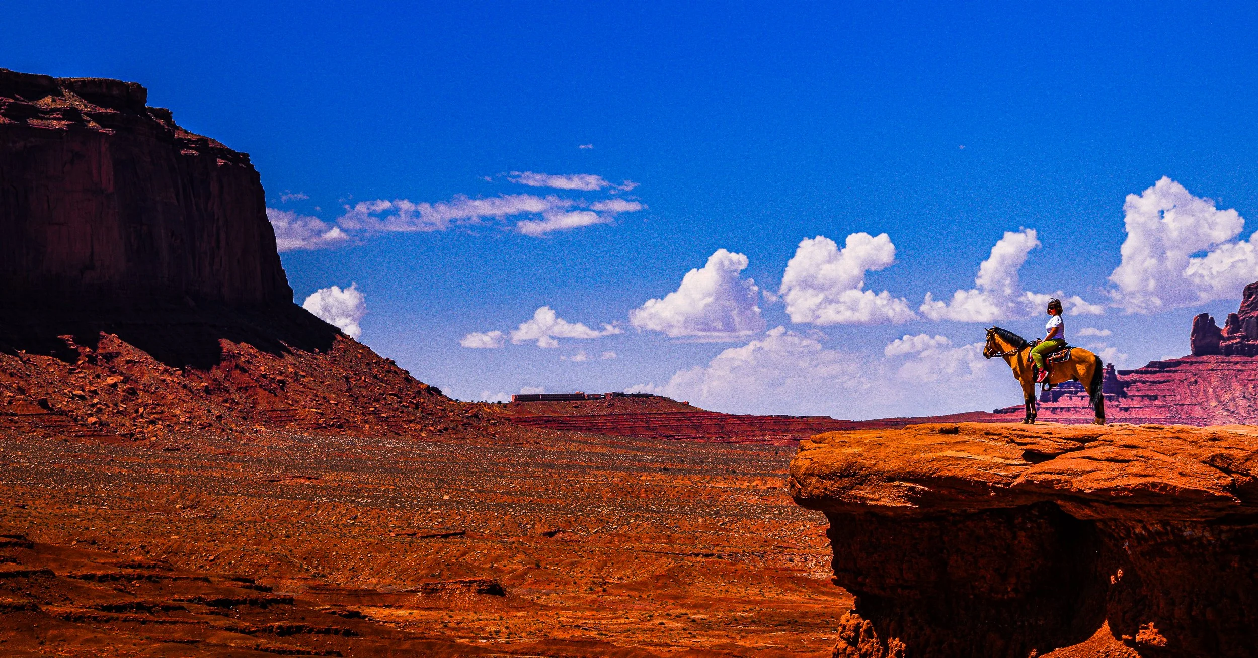 Rider on horseback overlooking a red-rock desert canyon beneath a blue sky with clouds