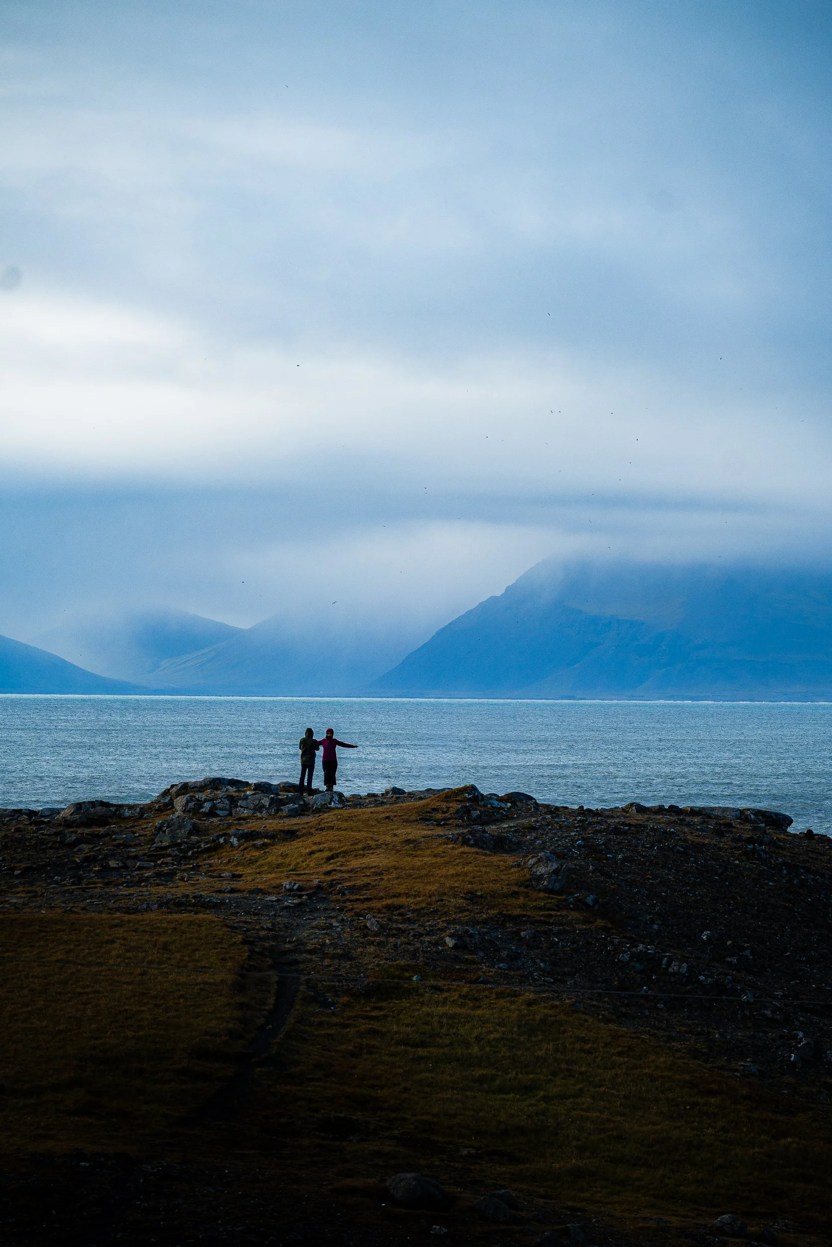 Two people stand on a rocky shoreline overlooking a calm blue fjord and misty mountains.
