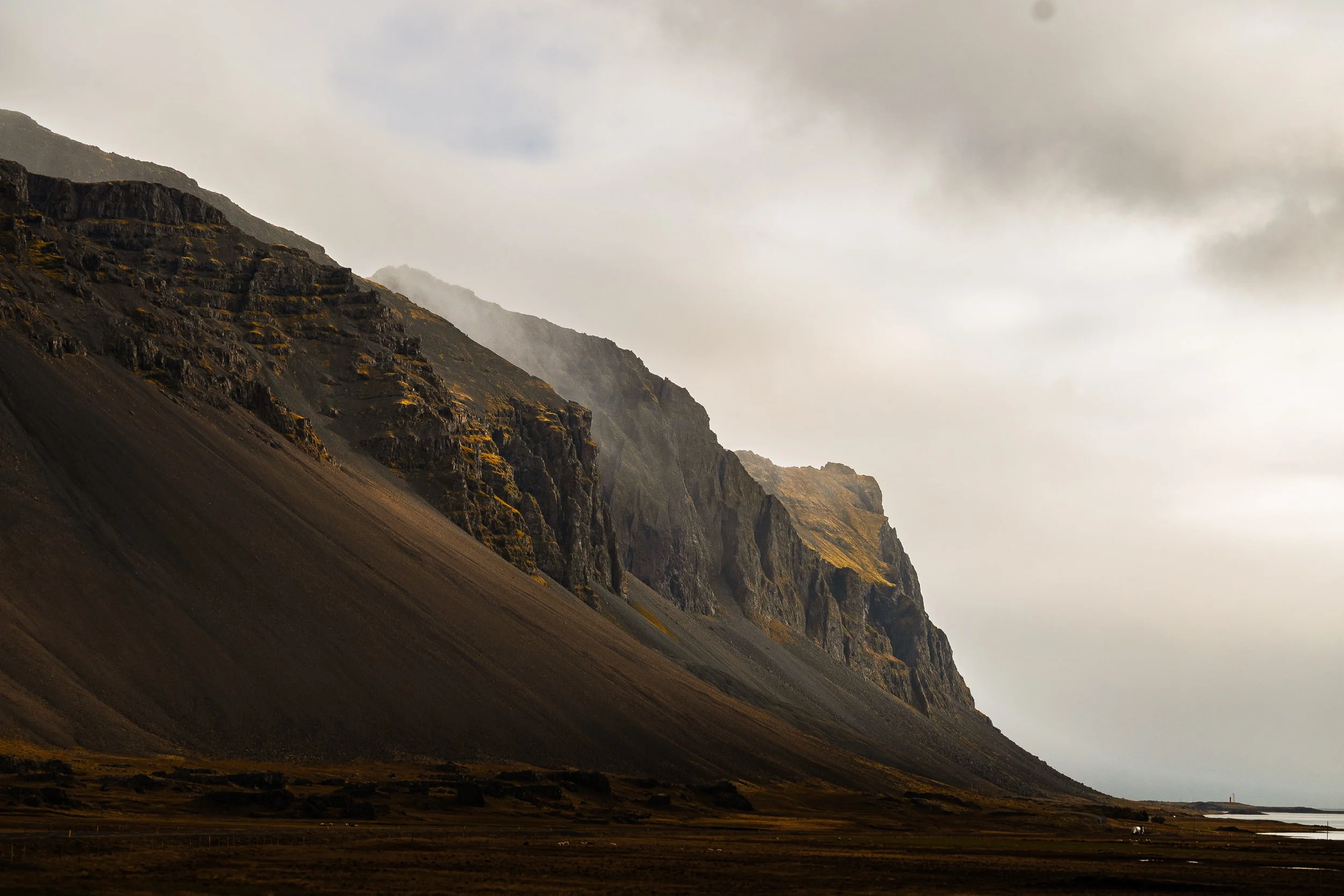 Dramatic Icelandic mountain cliffs beneath a cloudy sky, with a rugged valley and coastal water in the foreground