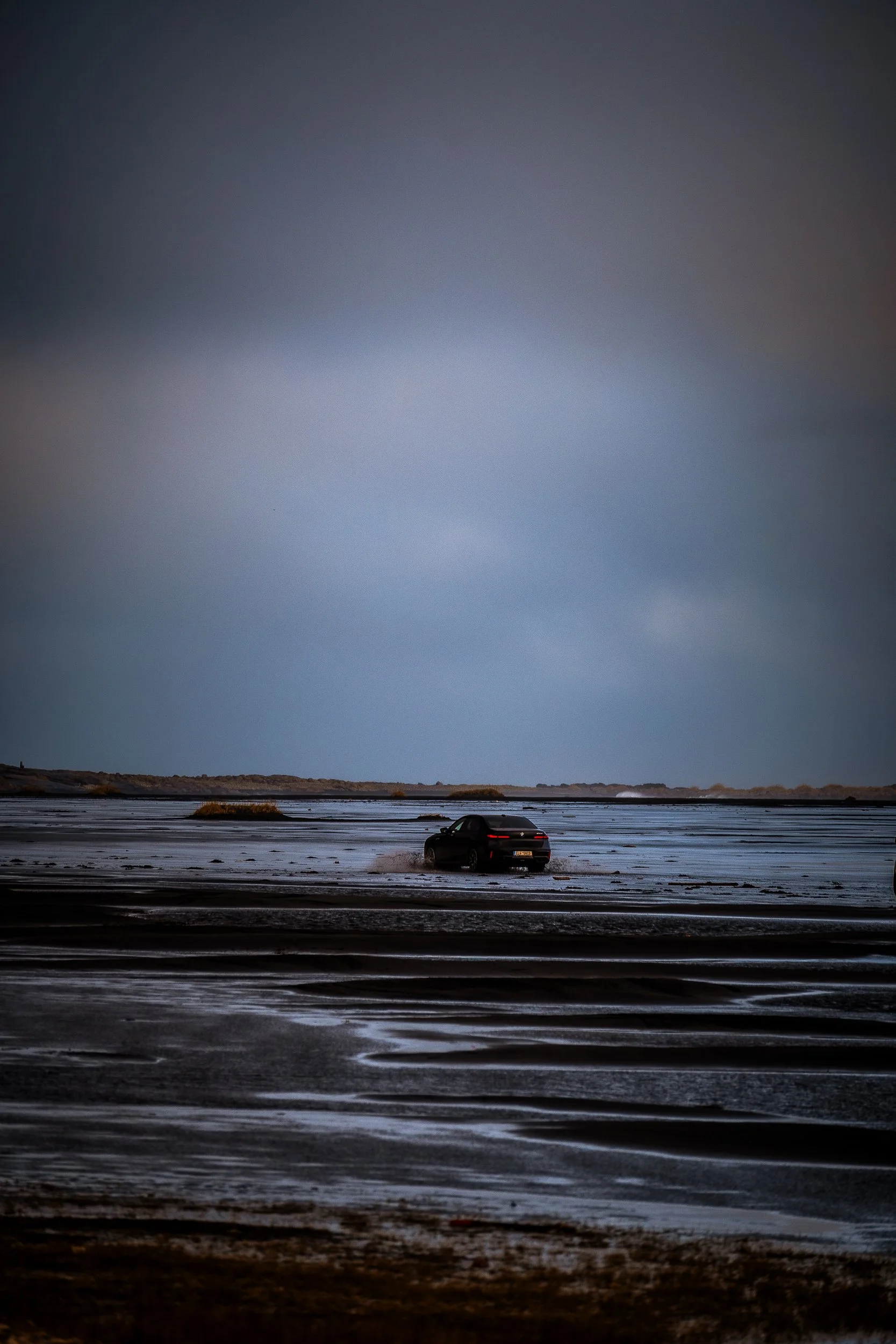 Black car driving across a wet tidal flat under a cloudy sky