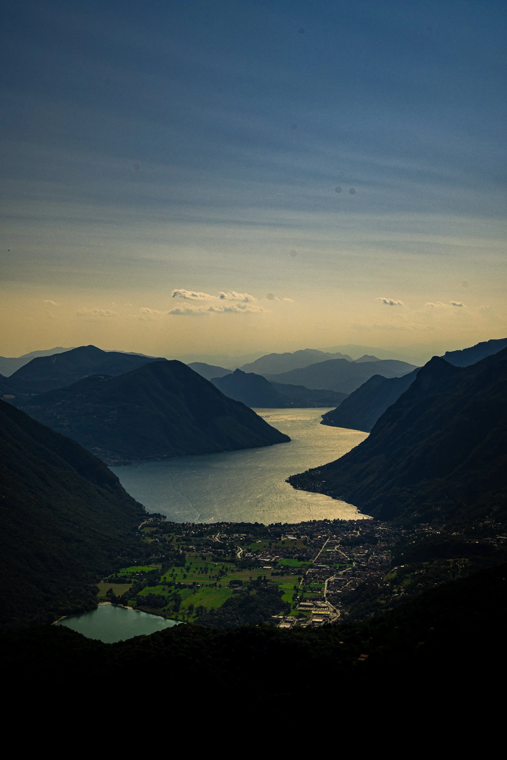 Aerial view of a sunlit alpine lake winding between dark mountain valleys, with a town and green farmland below at dusk