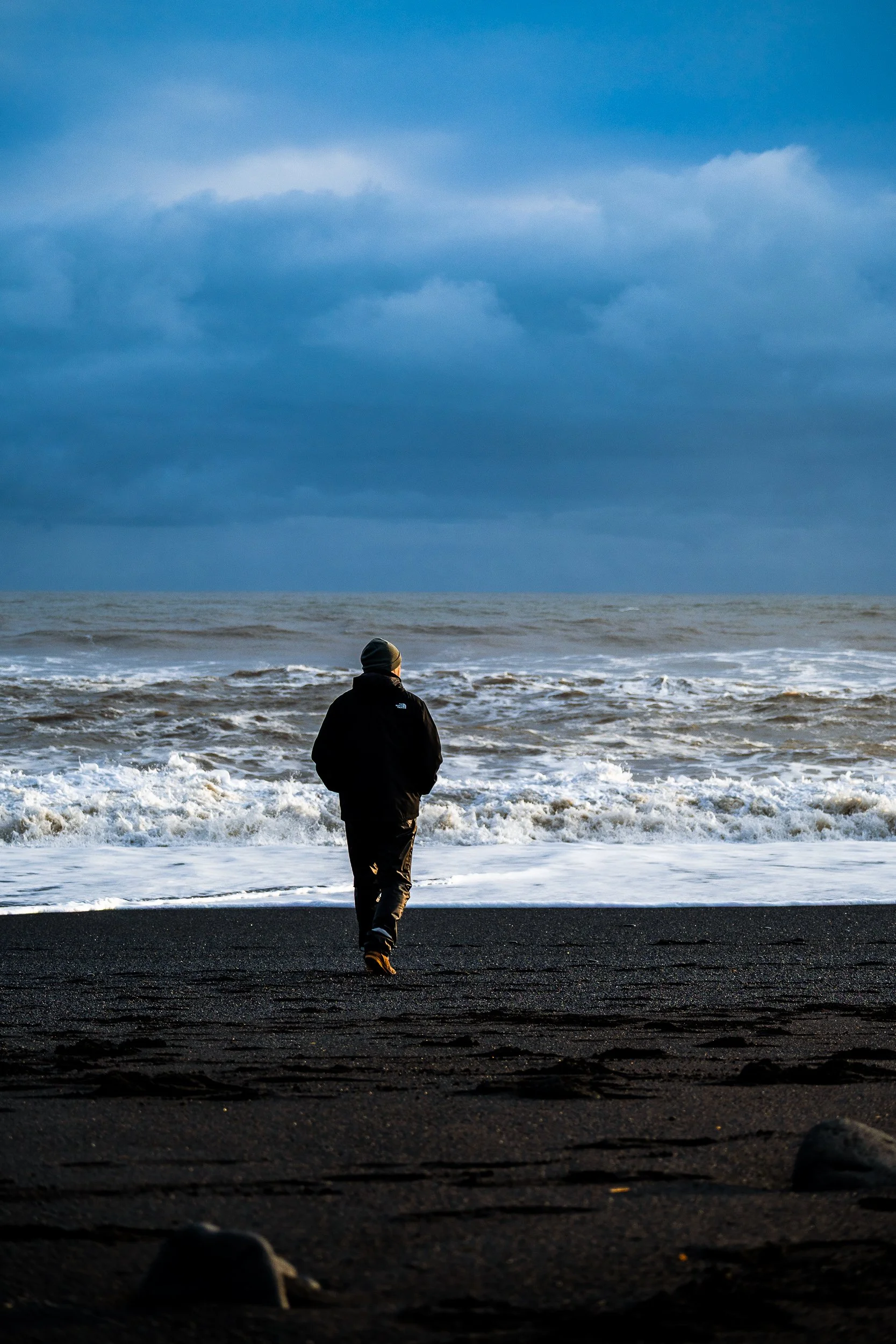Person in a black jacket and beanie walking toward waves on a black sand beach under a blue, cloudy sky