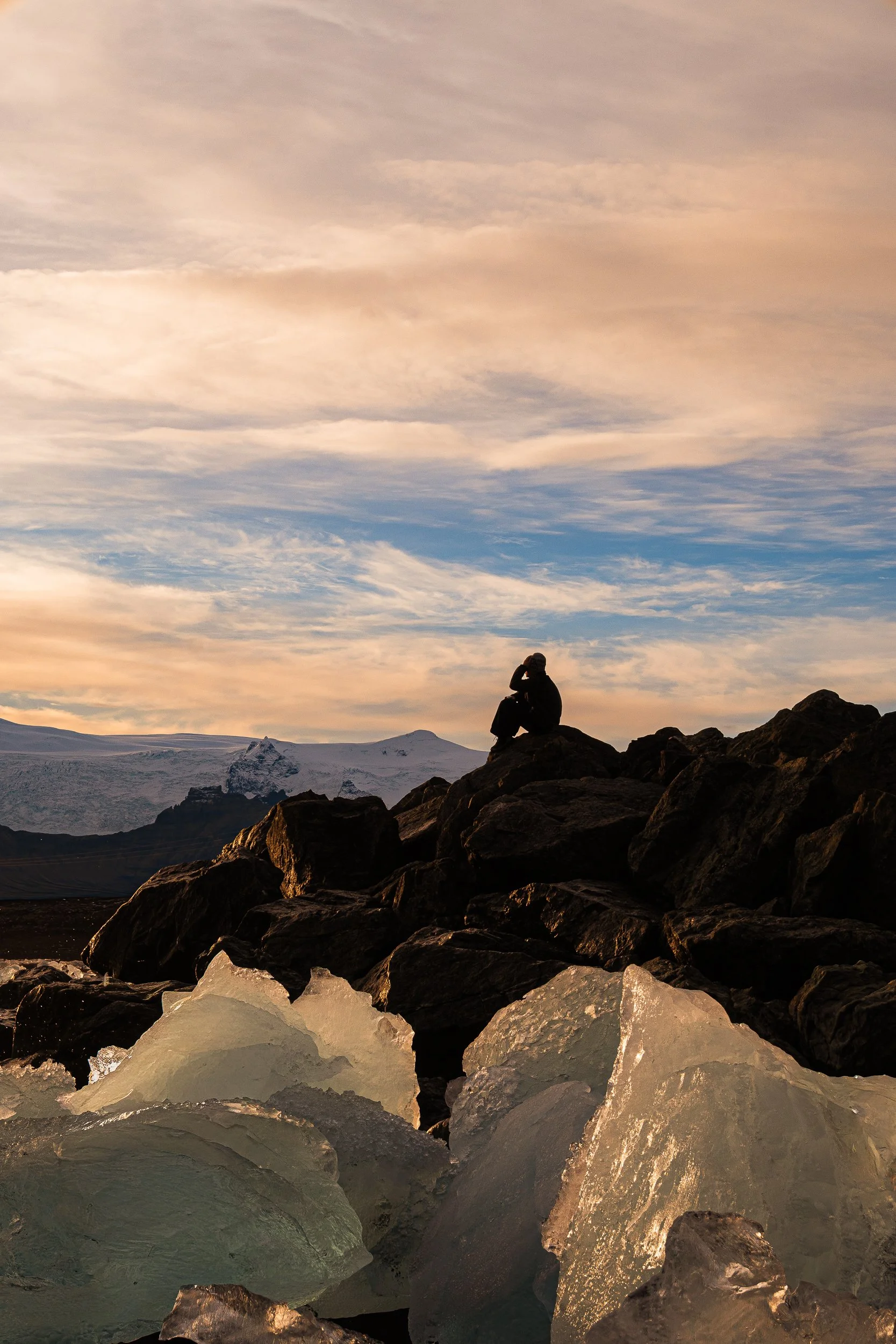 Silhouetted hiker sitting on rocky terrain overlooking a snow-covered mountain glacier at sunset