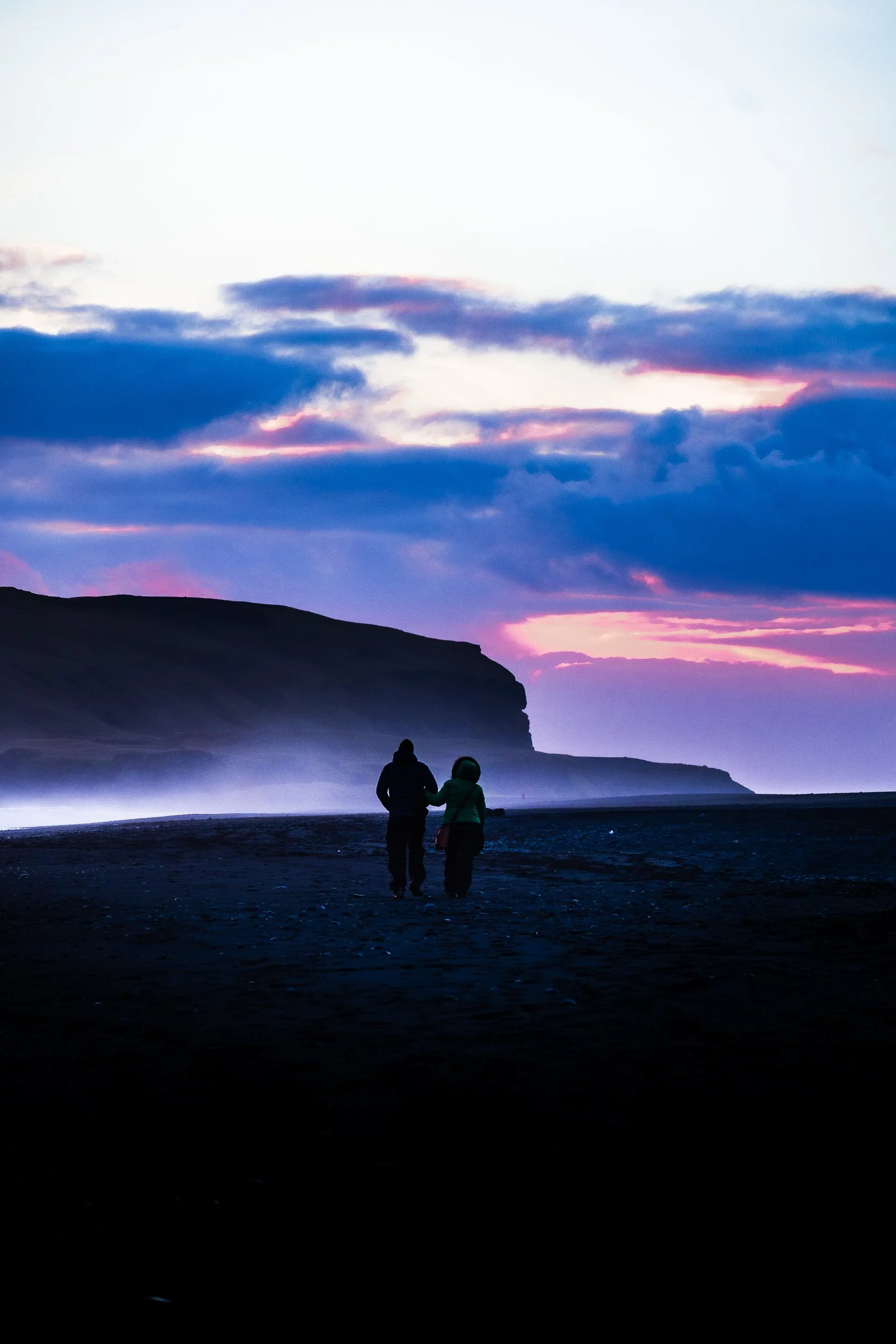 Two people walking across a dark beach beneath a colorful sunset sky and silhouetted rocky cliffs