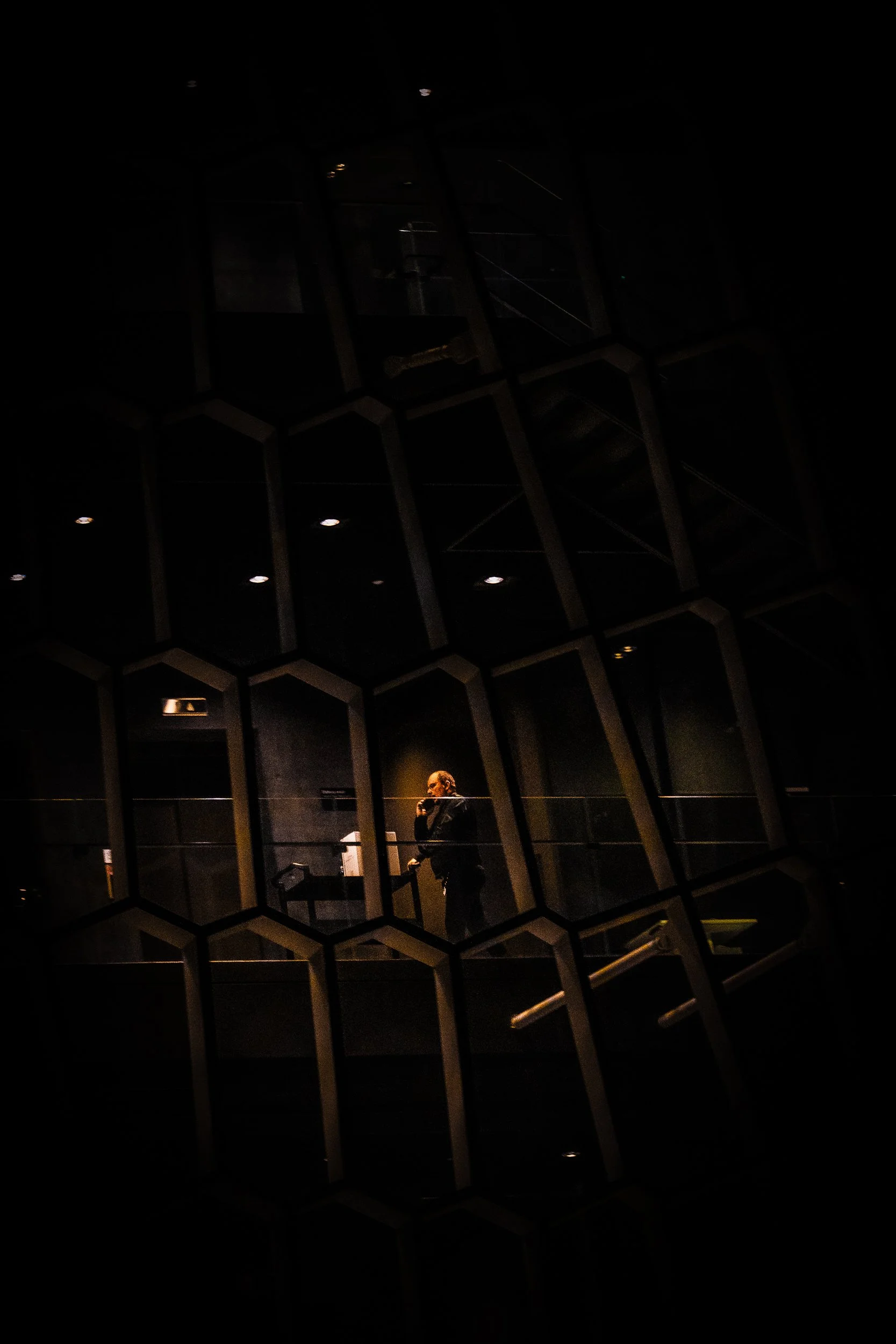 Dimly lit interior with geometric metal beams, ceiling lights, stair railings, and a person standing near an elevator or doorway.