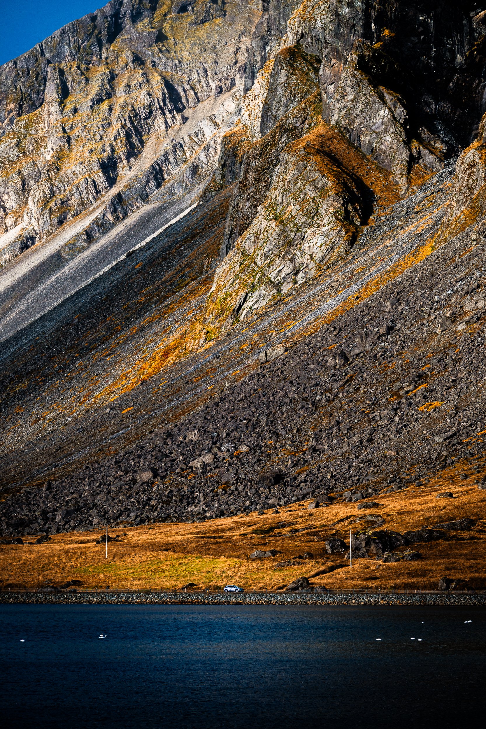 Dramatic jagged mountain cliffs above a rocky alpine slope, road, car, and dark blue lake with floating birds
