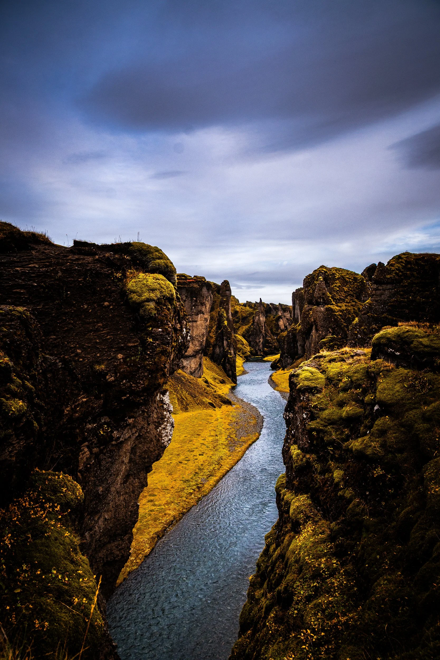 Moss-covered volcanic canyon with a winding river beneath a cloudy blue-gray sky