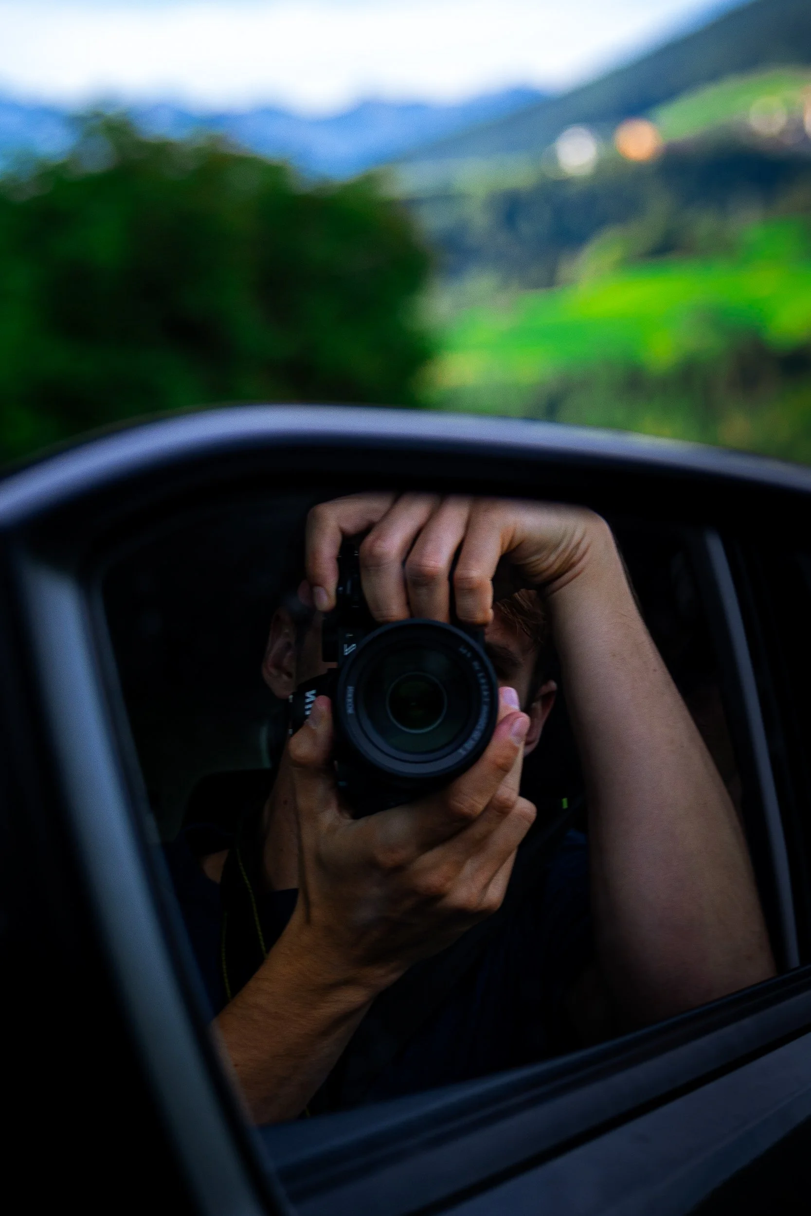 Photographer holding a camera while leaning out of a car window, with a blurred green mountain landscape in the background