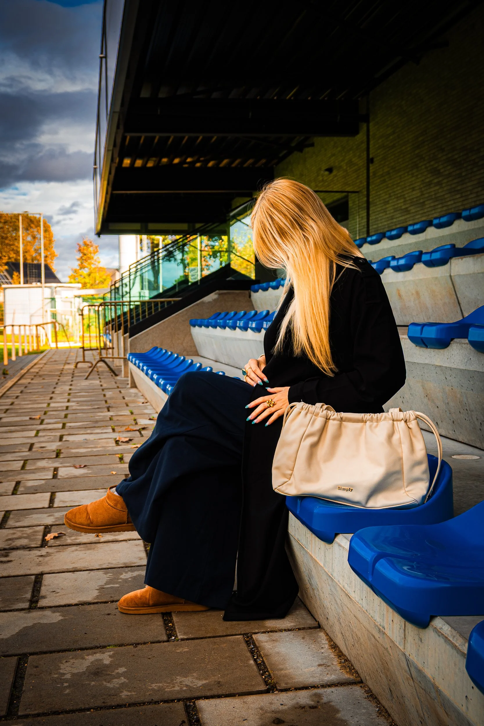 Woman in black clothing sitting in blue stadium seats with a beige handbag