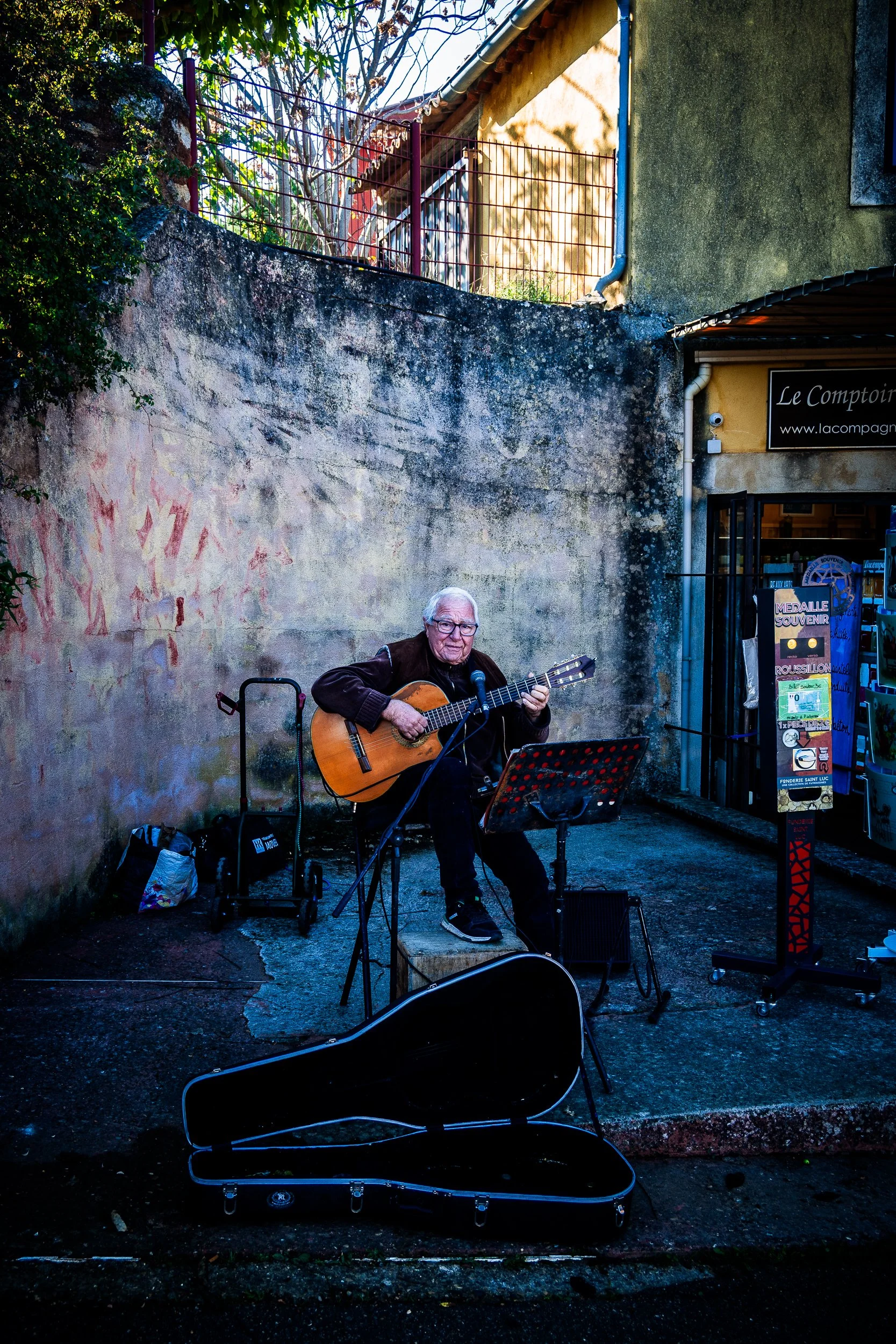 Street musician playing acoustic guitar outside a shop in a French village, with an open guitar case for tips.