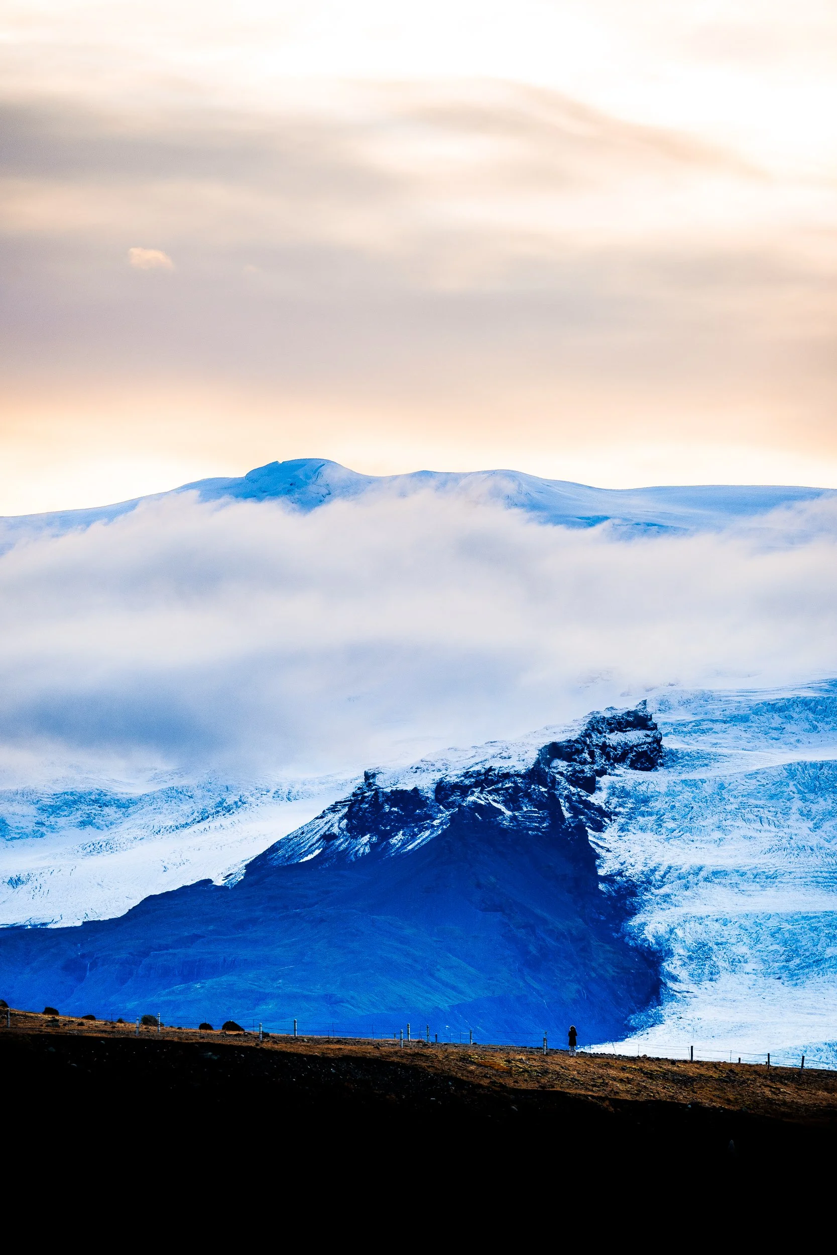 Snow-covered volcanic mountain and glacier beneath a pastel sky, with a lone hiker along a fenced ridge