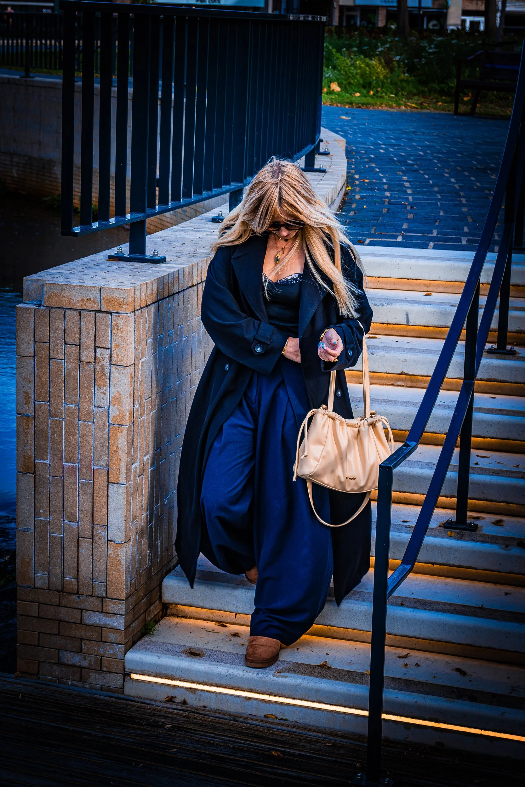 Blonde woman in a black coat and sunglasses walking down outdoor brick steps with a beige handbag