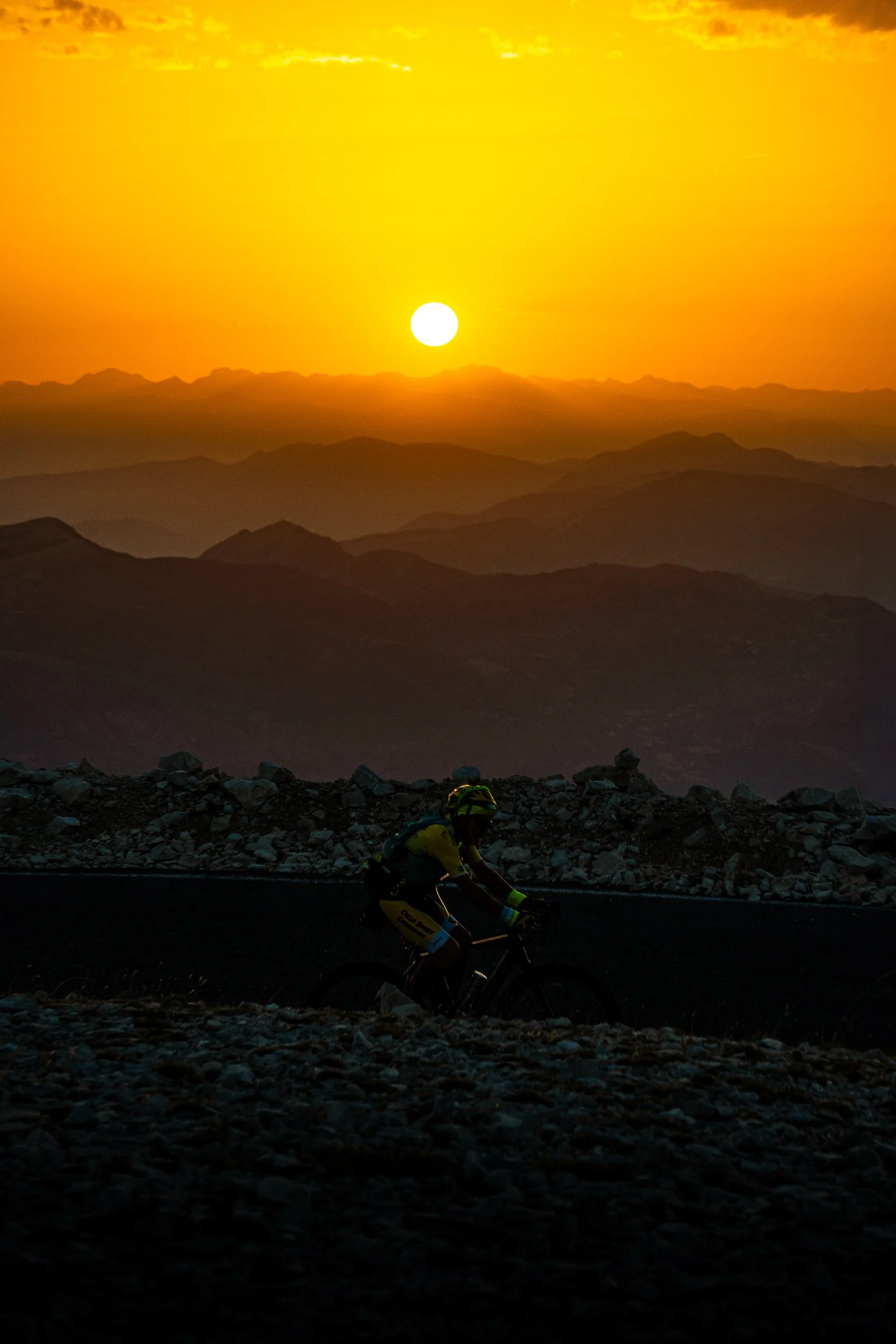 Mountain cyclist riding across a rocky landscape at sunset, with layered mountain silhouettes beneath a glowing orange sky.