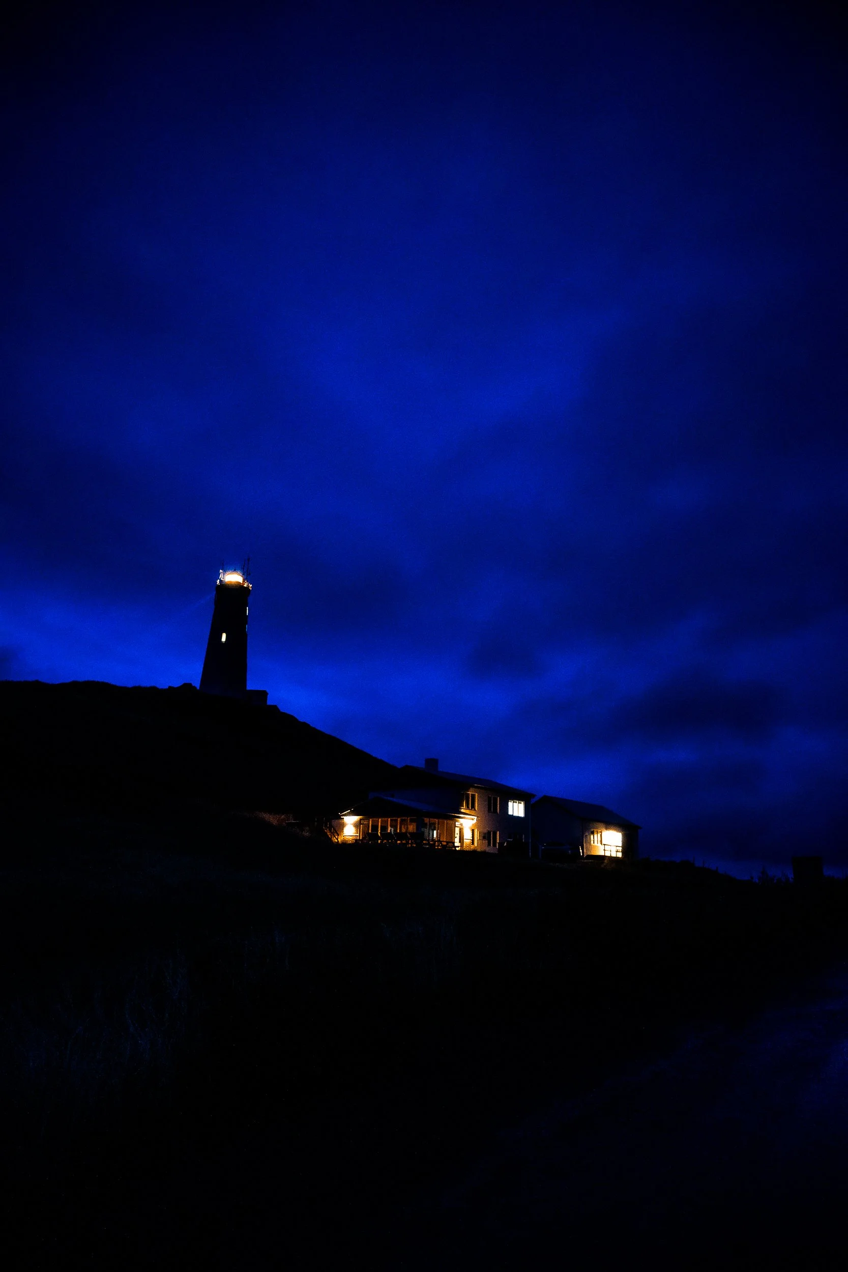 Lighthouse and illuminated coastal houses silhouetted against a deep blue twilight sky