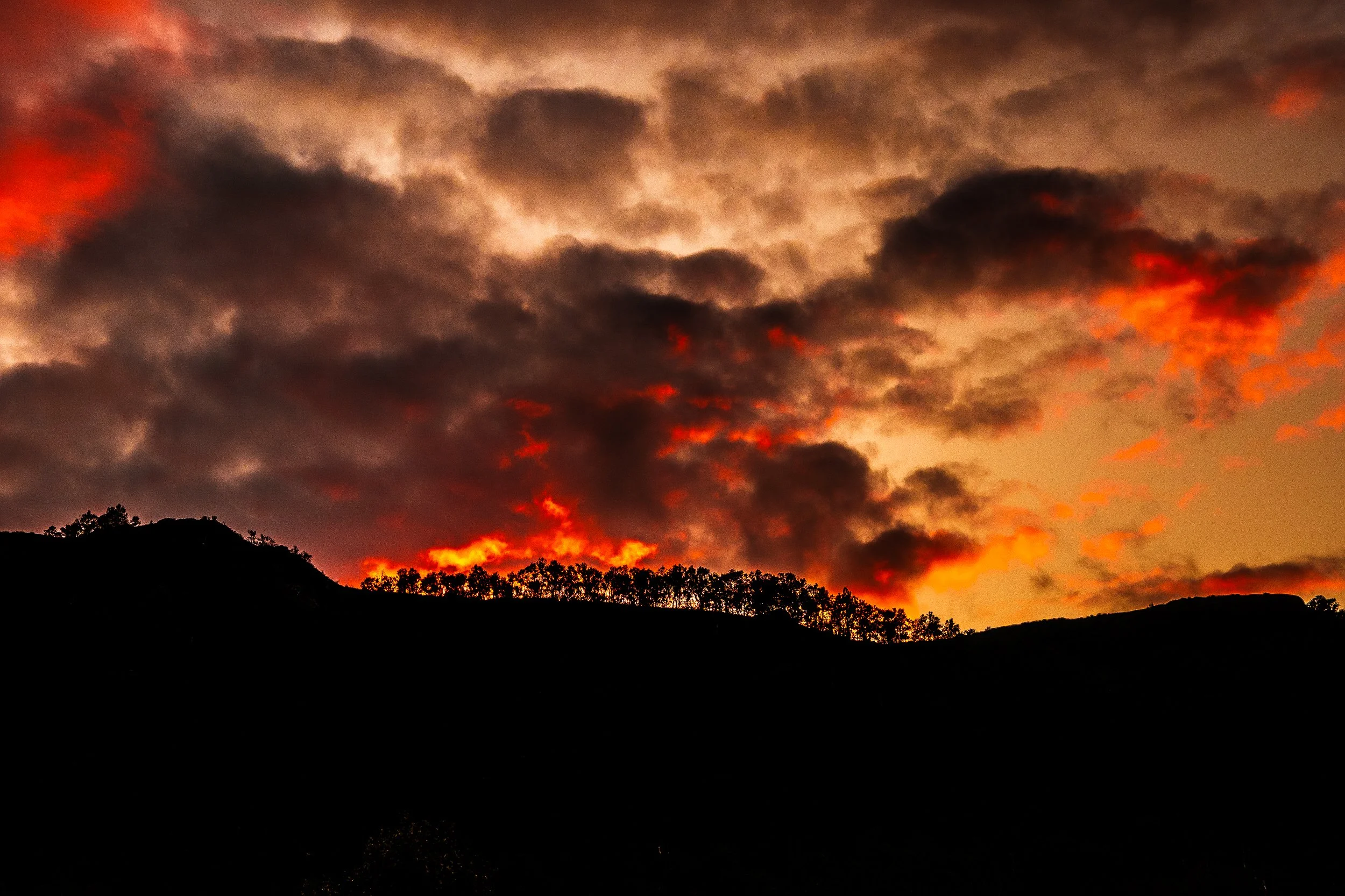 Dramatic orange sunset glowing through dark clouds above a silhouetted tree-lined hillside