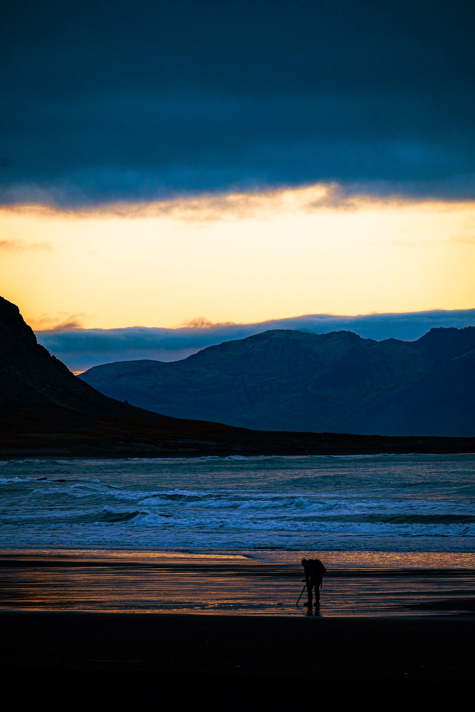 Silhouetted photographer standing on a beach at sunset with waves and rugged mountains in the background