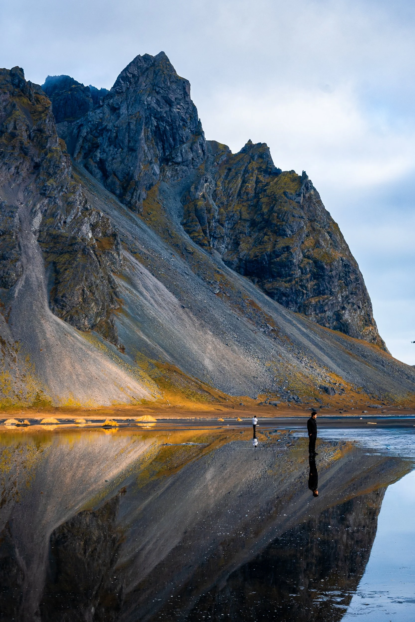 Two people stand beside a reflective tidal pool beneath rugged, jagged mountain cliffs.